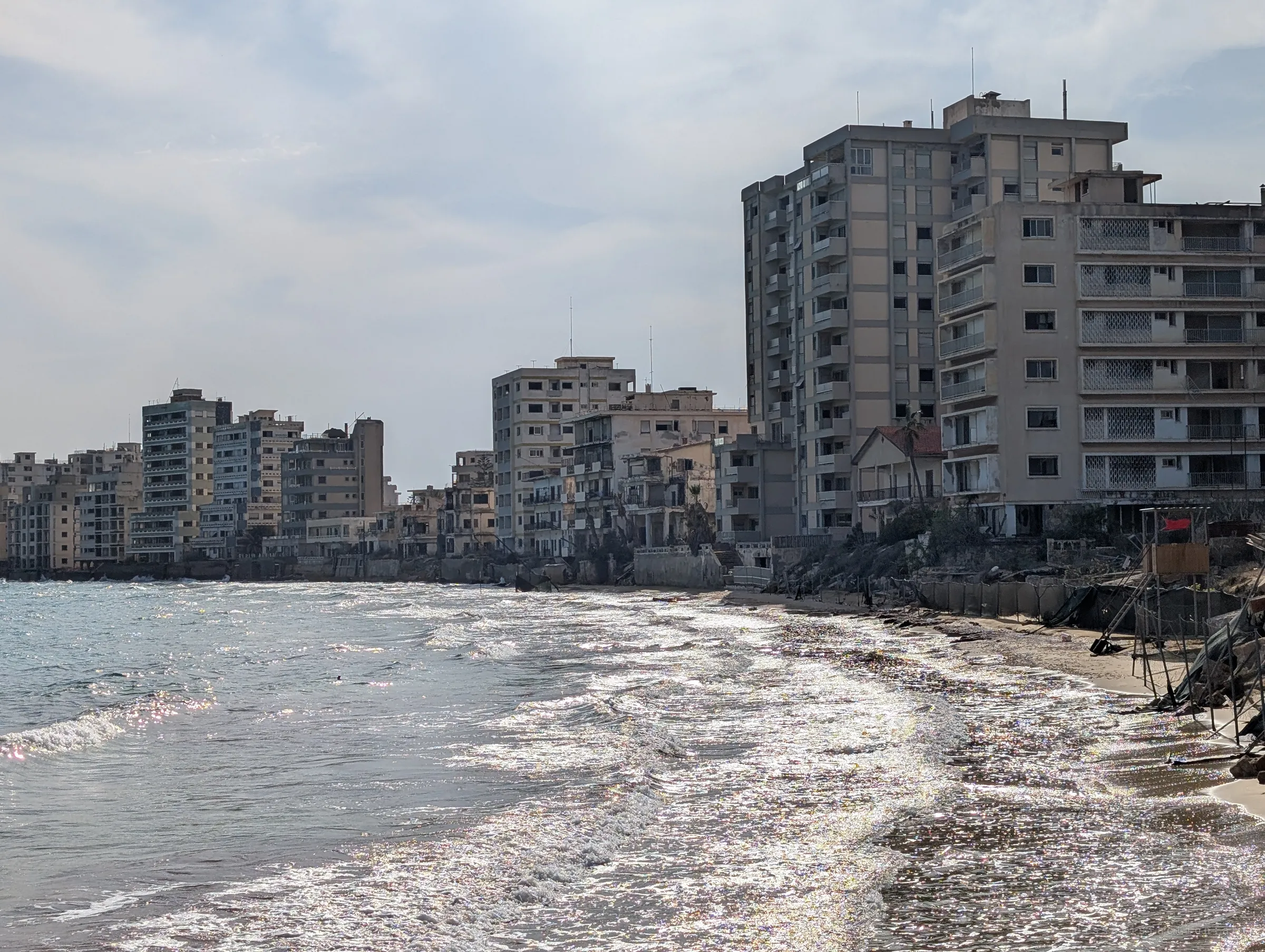 Waves glitter along an empty beach lined with a wall of abandoned high-rise hotels. Rusting fence posts and an old lifeguard tower mark where the shore was sealed off.