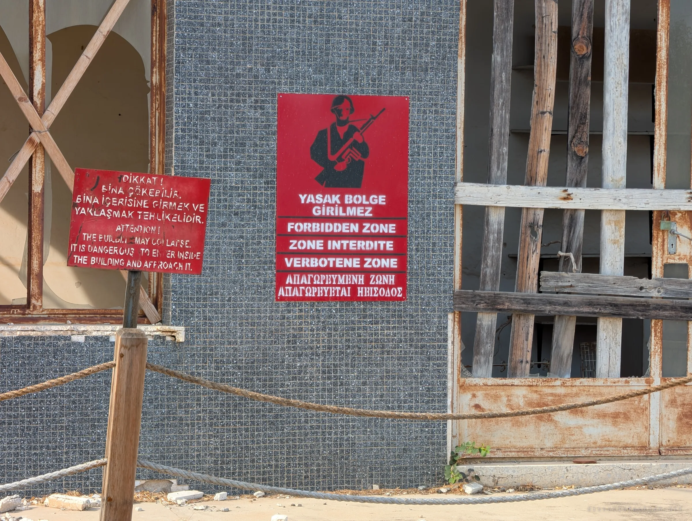 Red multilingual 'forbidden zone' sign with a soldier silhouette beside a collapse warning, fixed to a mosaic-tiled wall between boarded, rusted doorways. The roped-off frontage shows how the abandoned buildings are sealed off.