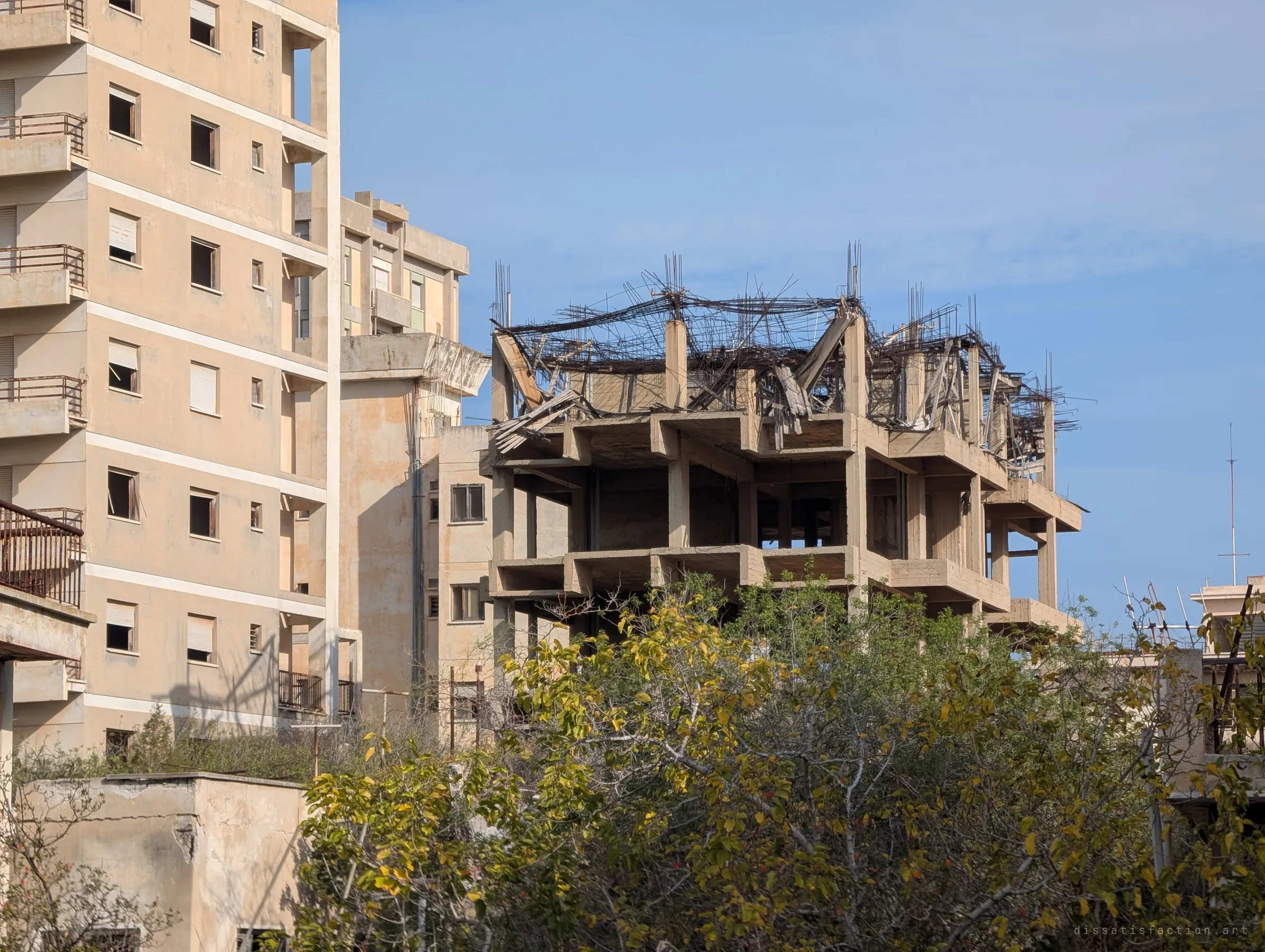 Empty sun-bleached apartment blocks stand behind an unfinished concrete frame crowned with a tangle of rusted rebar, trees growing up in front. Decades of neglect left construction frozen mid-build.