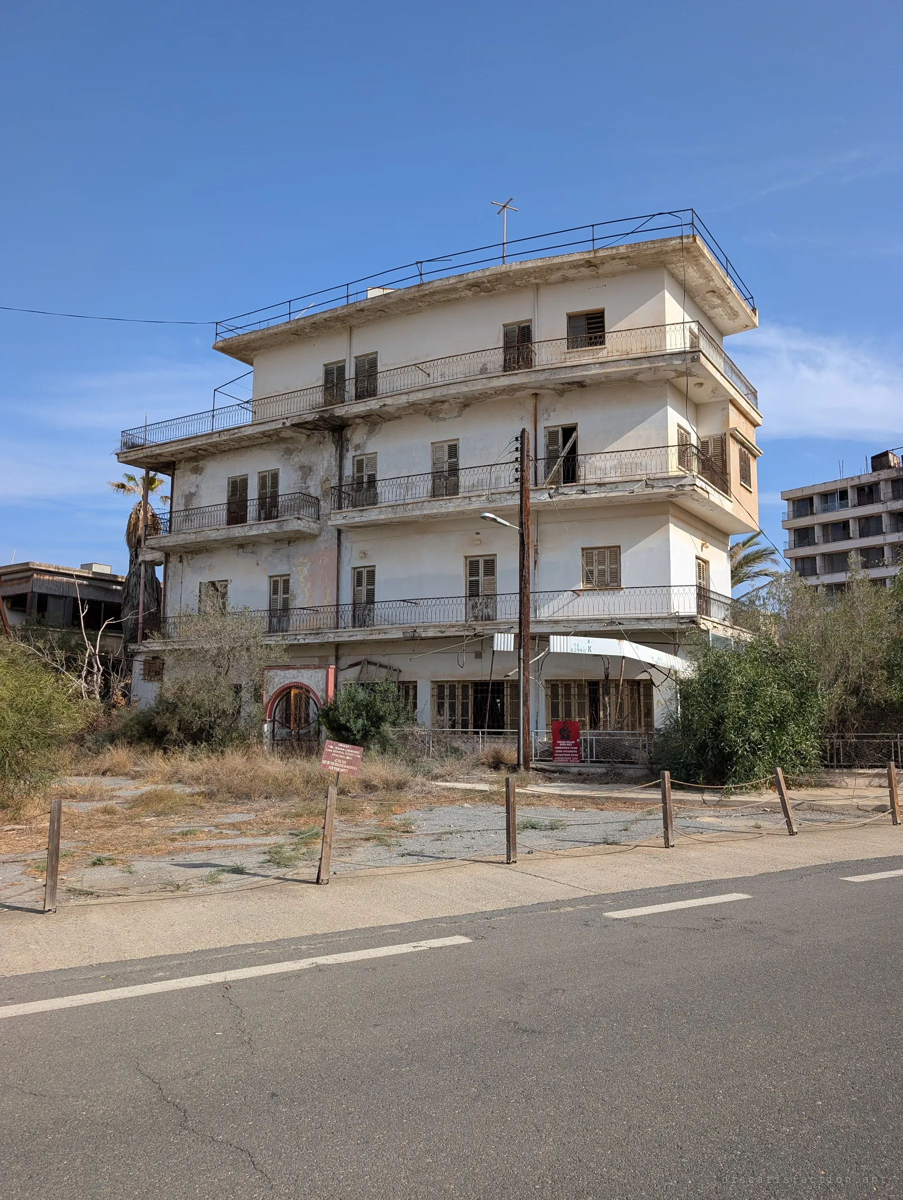 A four-storey shuttered villa with wrought-iron balconies and a rooftop cross stands behind a roped-off verge, a faded shop awning above its ground floor. Across the empty road, weeds have taken the forecourt.