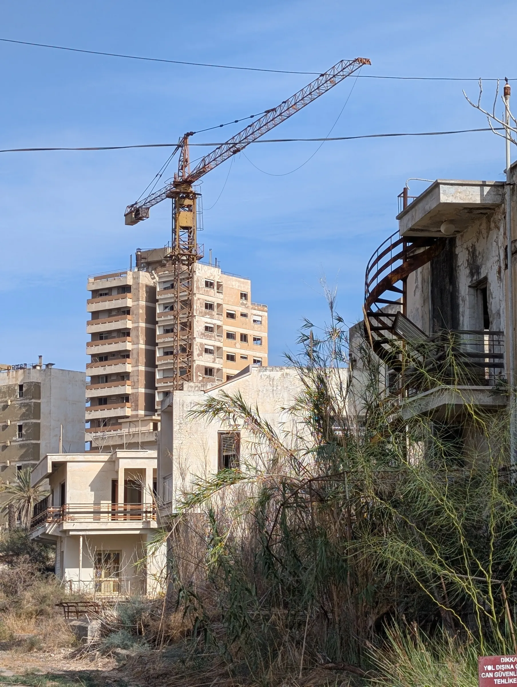 A rusted tower crane still stands over an empty high-rise, framed by overgrown villas and a corroded spiral staircase. Work stopped here and never resumed.