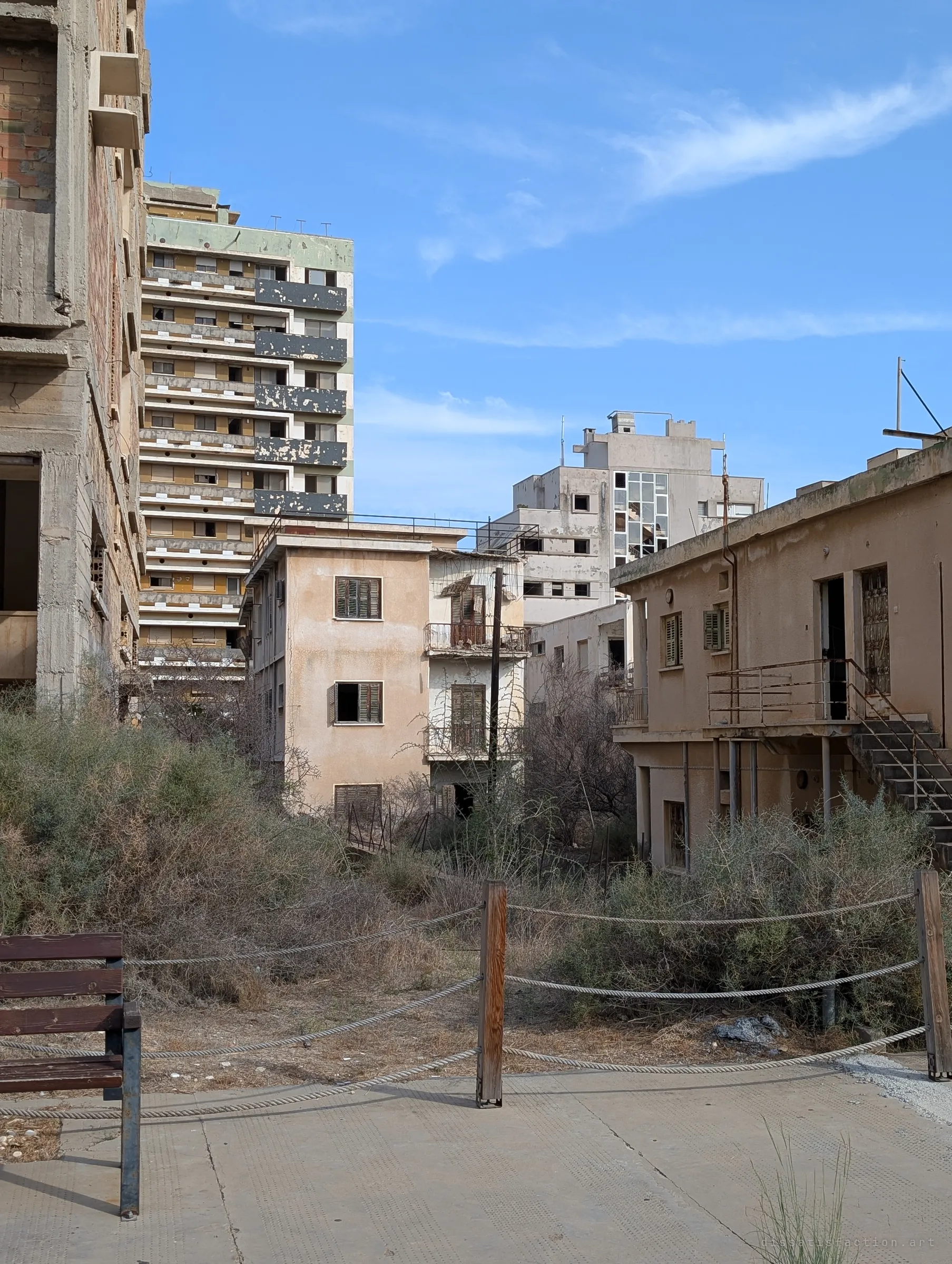 A roped-off path and bench face a lane of overgrown houses, with gutted high-rise blocks rising behind them. Scrub has filled the gaps between buildings.