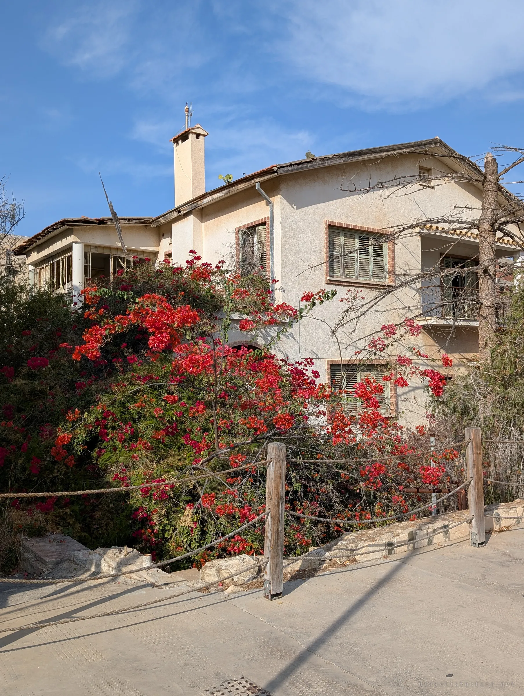 Bright red bougainvillea spills over the rope fence in front of a shuttered two-storey house with brick-framed windows and a sagging roof. The garden has outlived its owners.