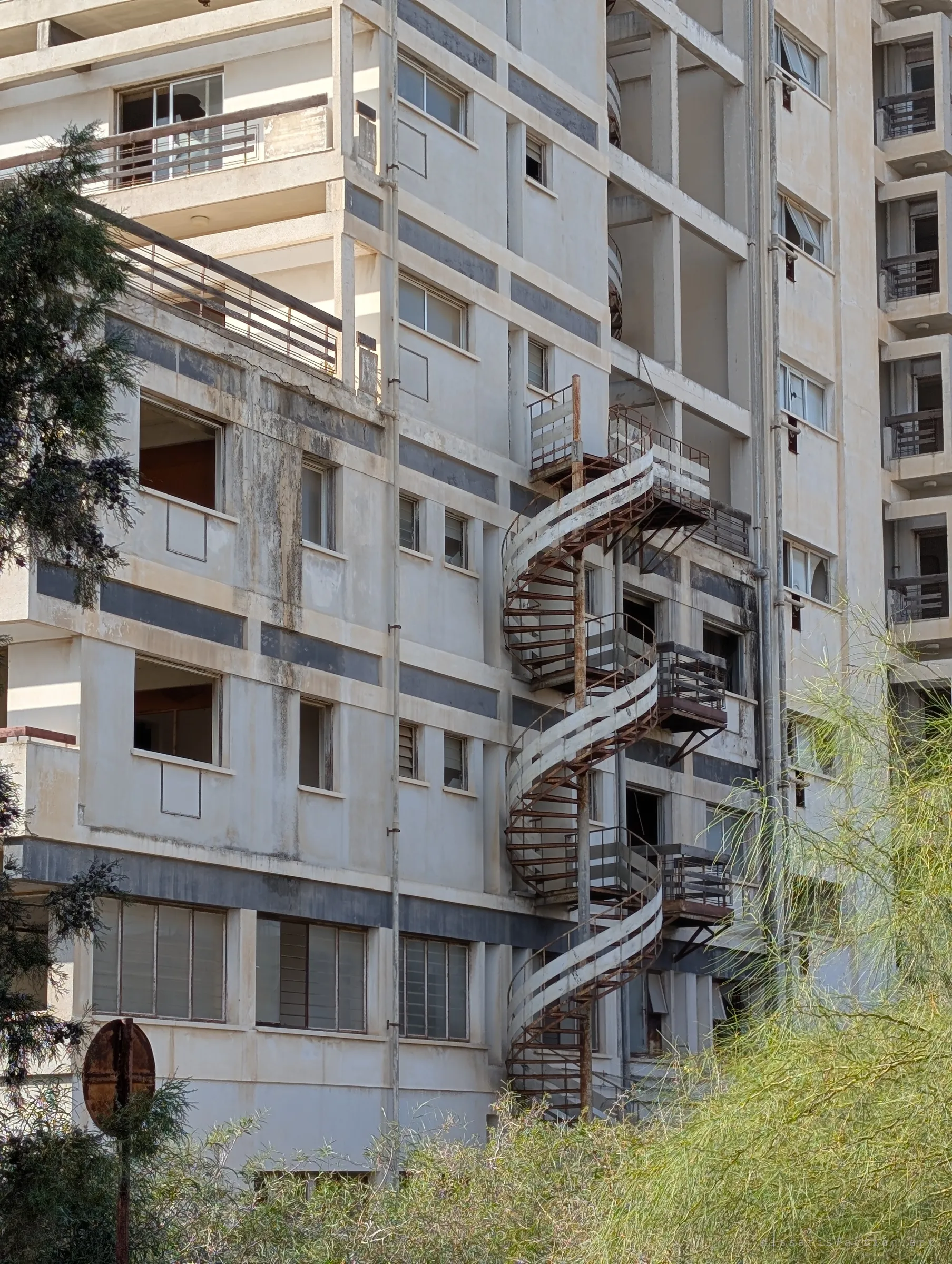 A rusted spiral fire staircase twists up the stained facade of an abandoned hotel block, its window frames empty. Greenery creeps up from below.