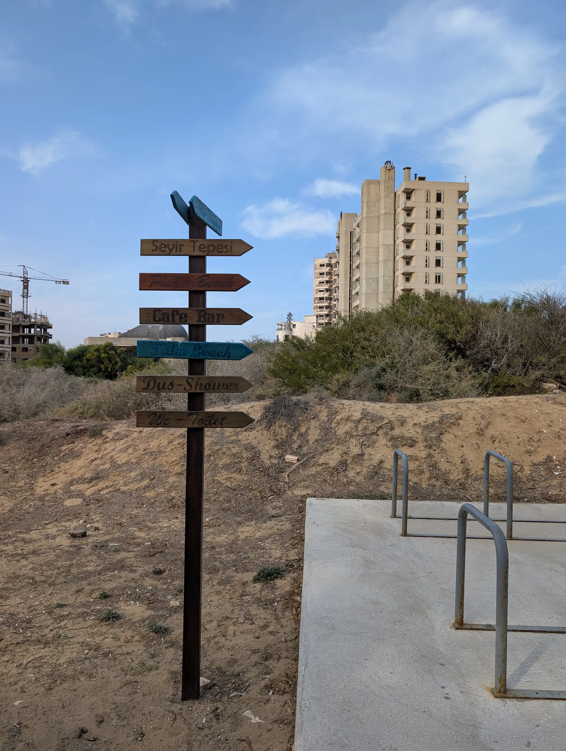 A wooden signpost of painted arrows points toward the beach, a café and showers on a sandy dune, with an abandoned hotel tower rising behind the scrub. The visitor path now runs past a town nobody lives in.
