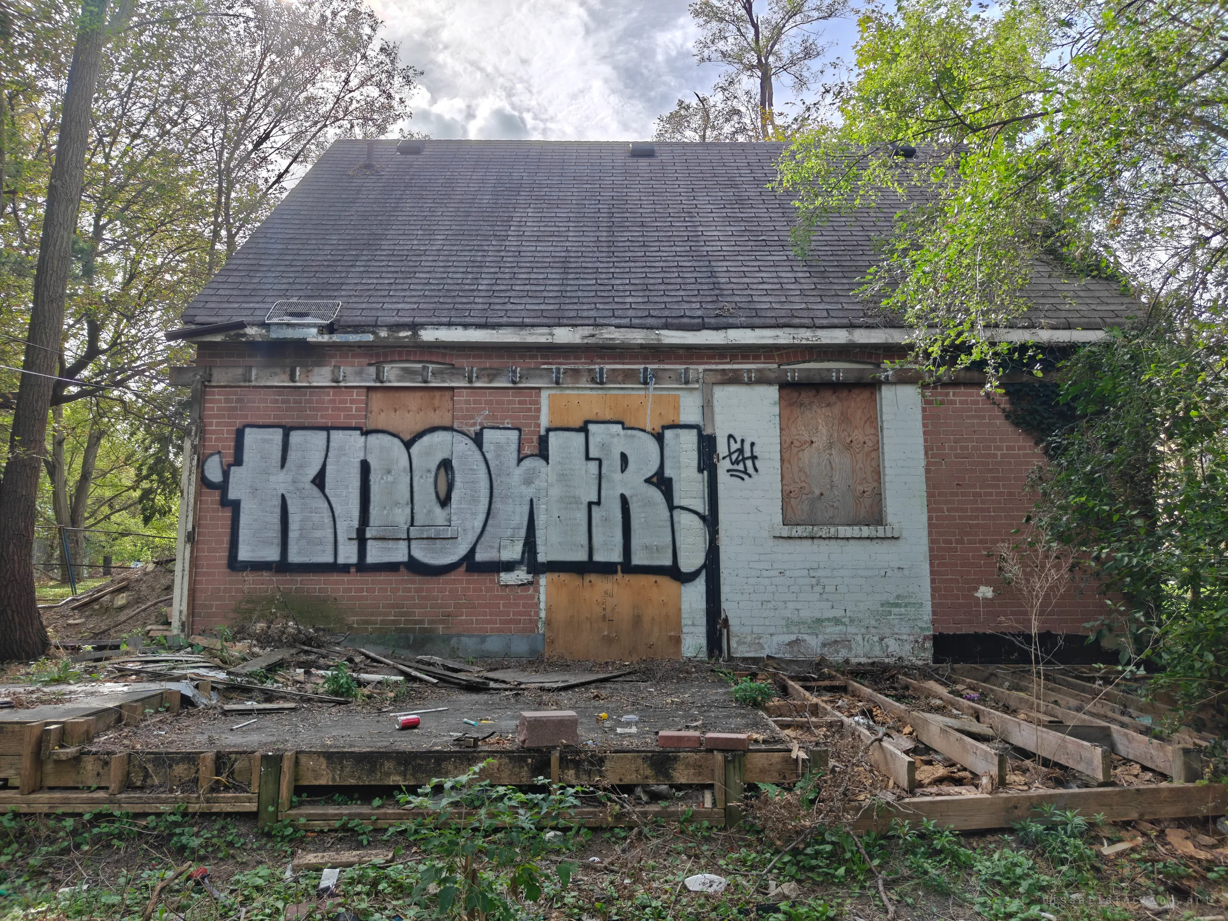 The back of a small abandoned brick house, doors and windows boarded with plywood and a large silver graffiti piece across the wall. Its rotted deck frame lies open in the overgrown yard, setting the scene before going inside.