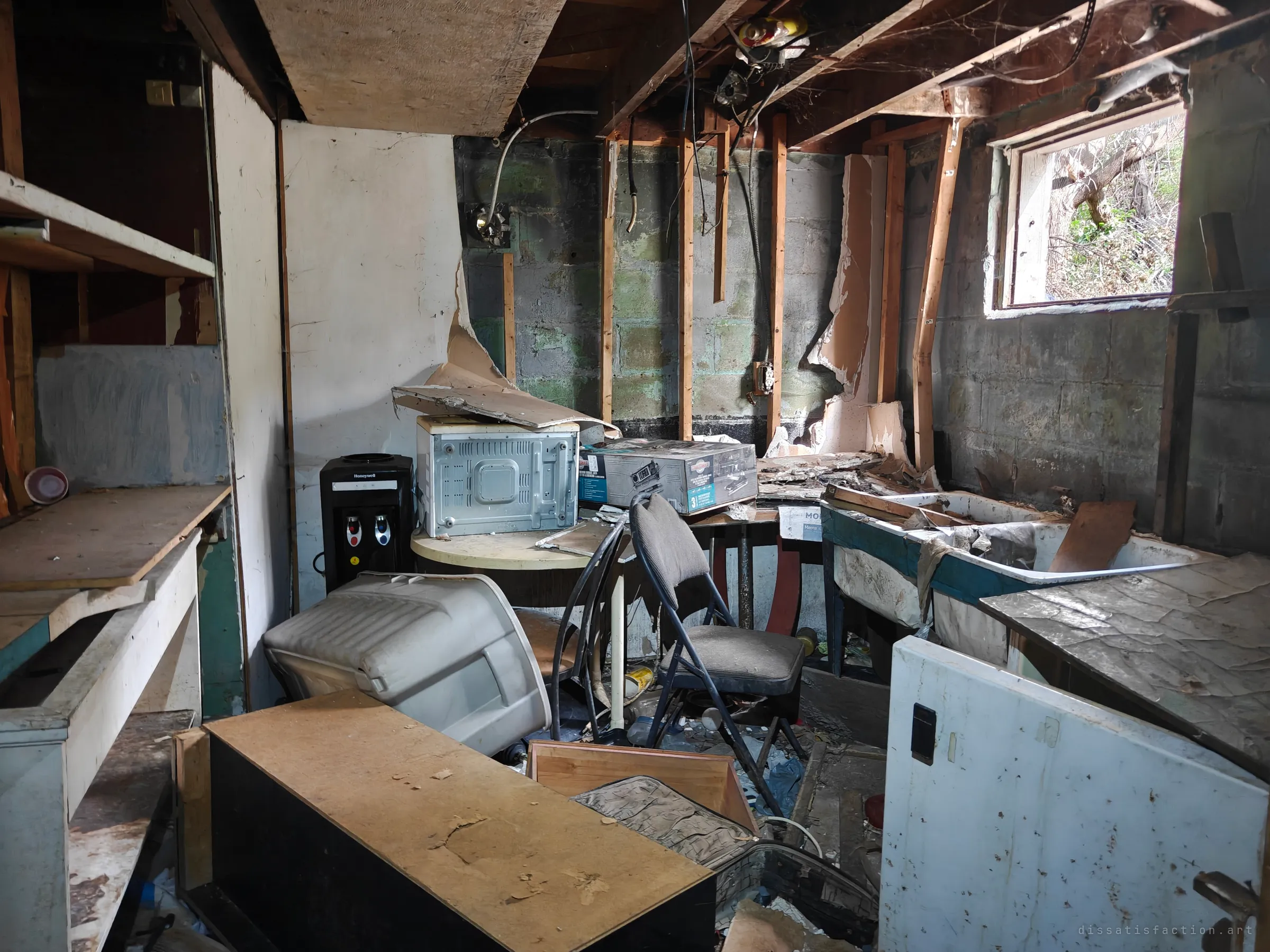A gutted basement crammed with a water cooler, a microwave, a folding chair and a cracked laundry sink under exposed joists and dangling wires. Daylight from a small window shows only tangled brush outside.