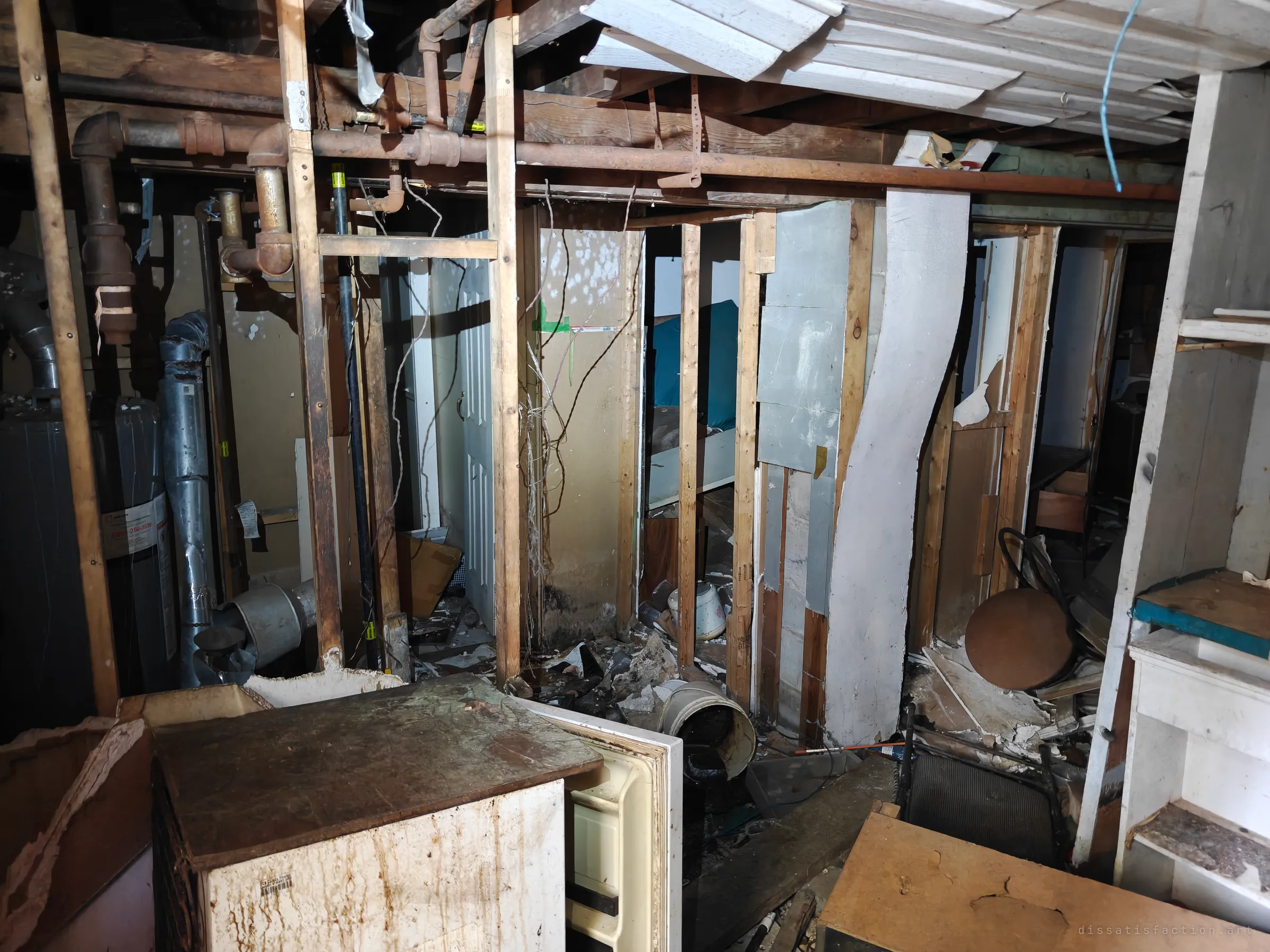 Stripped basement framing with rusted pipes overhead, a water heater on the left and a chest freezer and debris in the foreground. Open stud walls let you see straight through the old partitioned rooms.