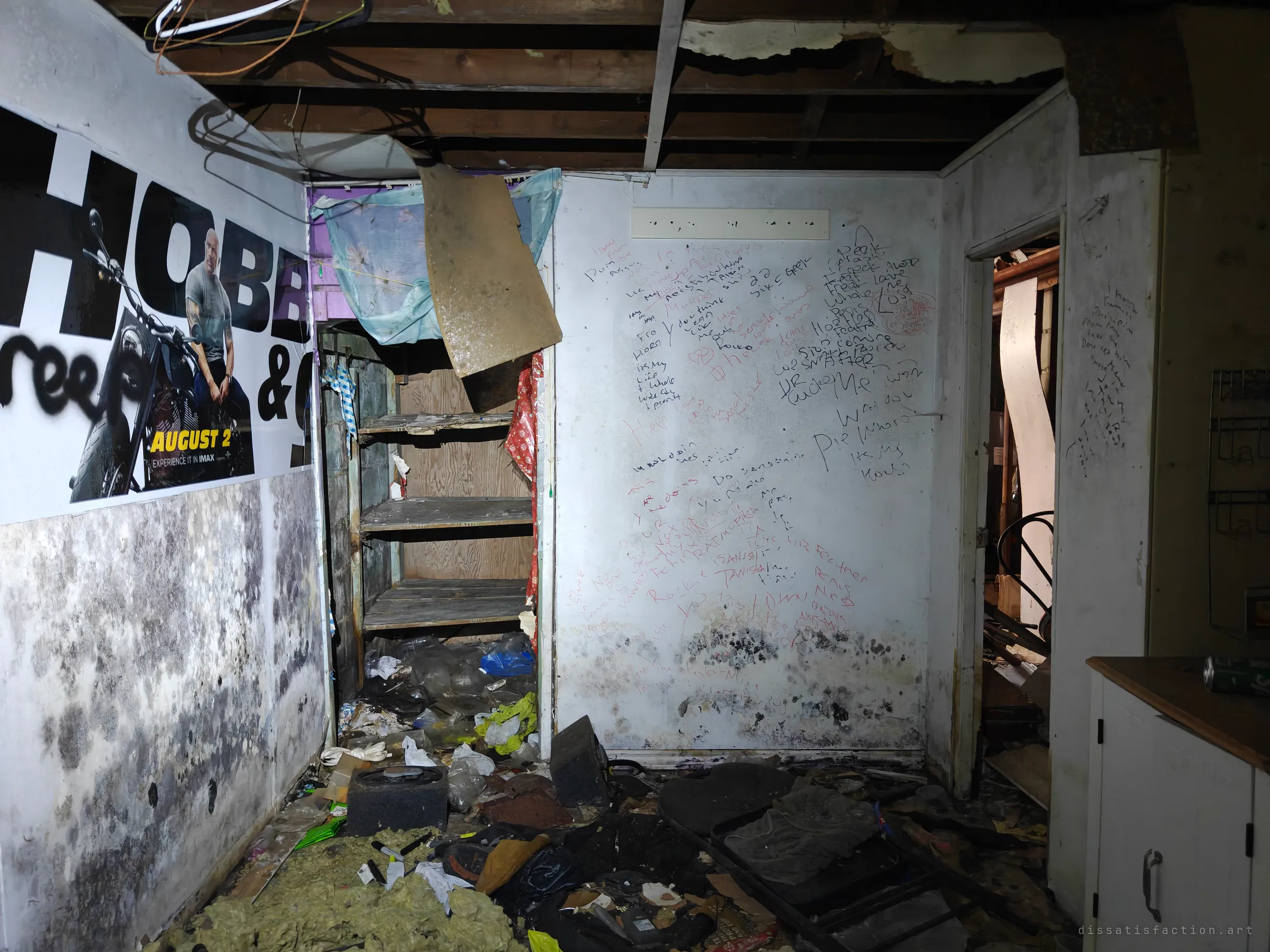 A mould-streaked room where a movie poster peels beside a wall covered in handwritten messages left by visitors. Trash, insulation and an old speaker box litter the floor.