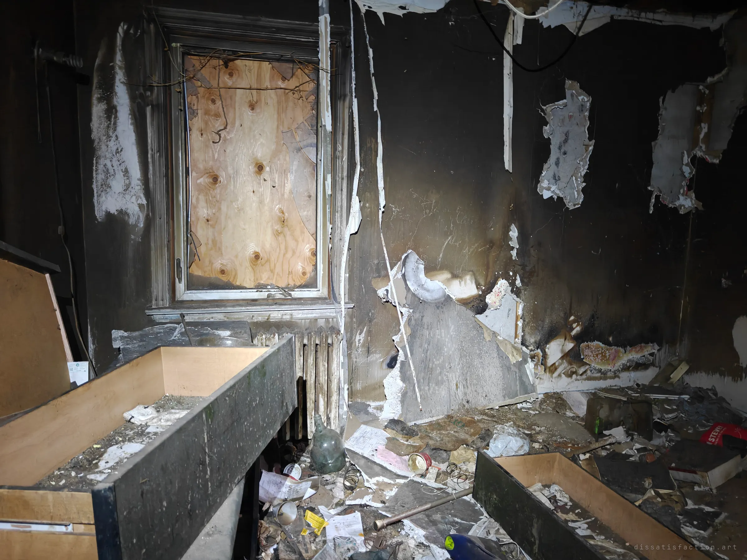 A fire-blackened room with a boarded window above an old radiator and wallpaper hanging in scorched strips. Pulled-out drawers and burnt debris, a green glass jug among them, cover the floor.