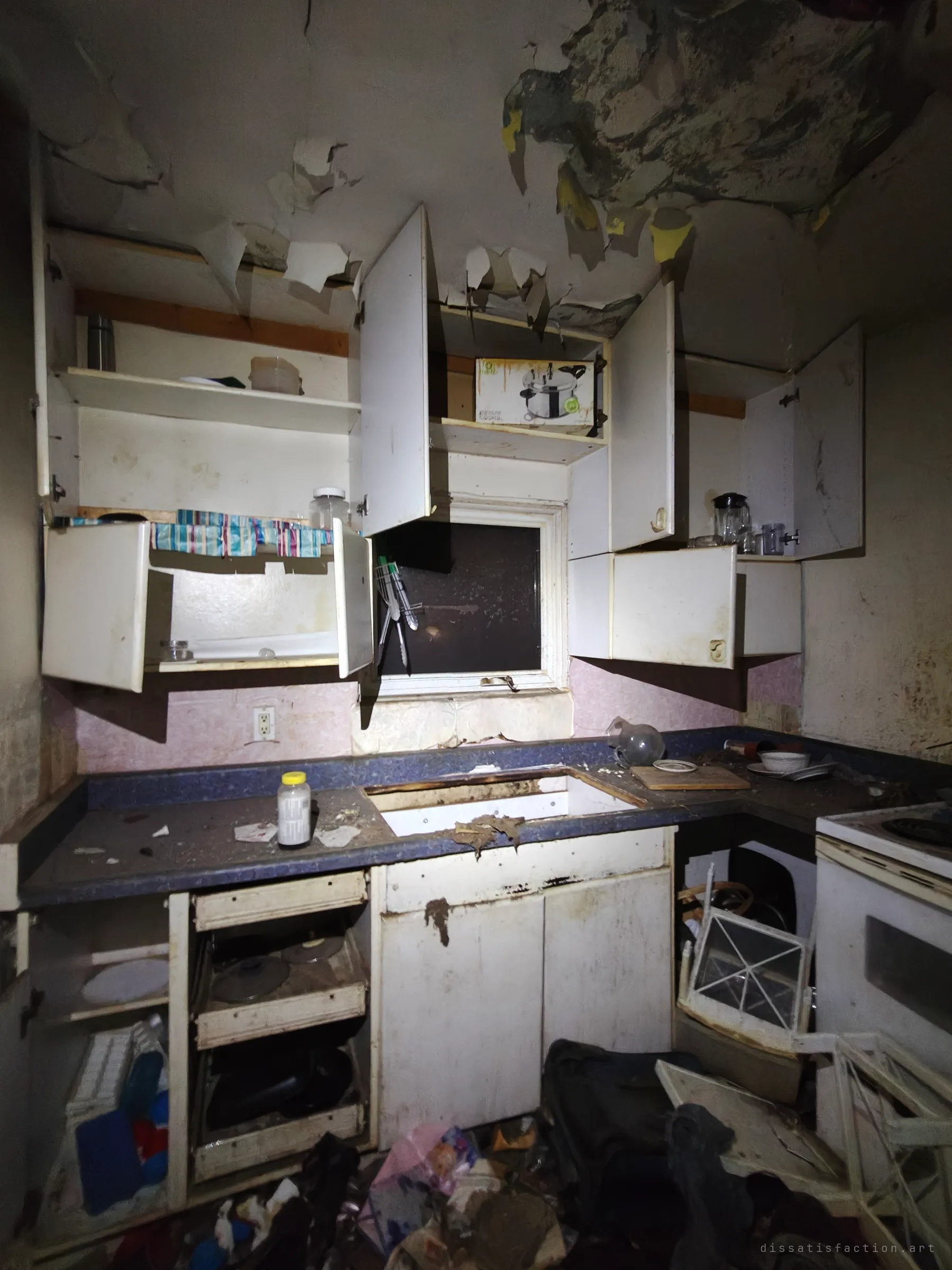 A small kitchen with every cabinet door hanging open, the sink ripped out of the counter and the ceiling paint curling away in sheets. A pressure-cooker box still sits on a shelf above the dark window.