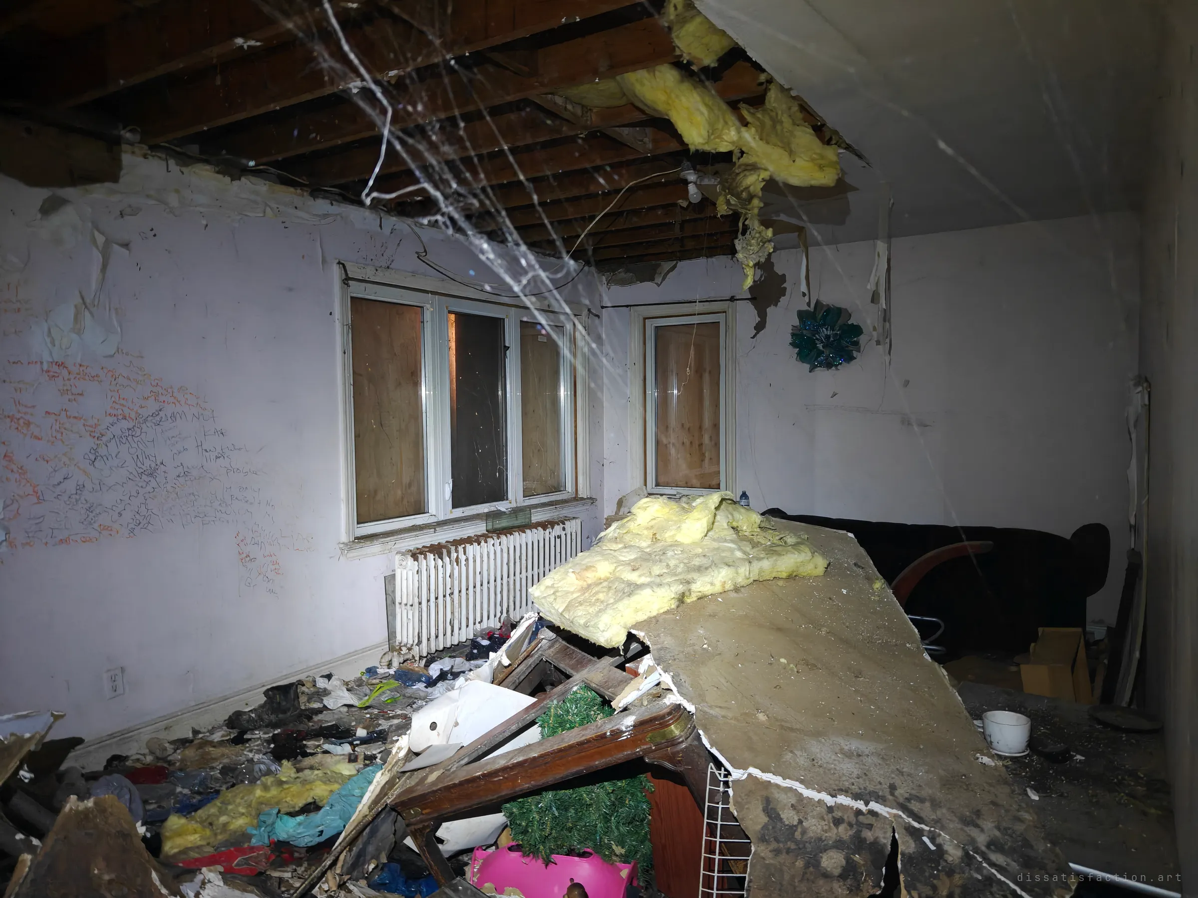 A living room where the ceiling has caved in, dumping insulation and plaster onto a toppled cabinet. Boarded windows, a radiator and a wall of scrawled messages sit behind a curtain of cobwebs.