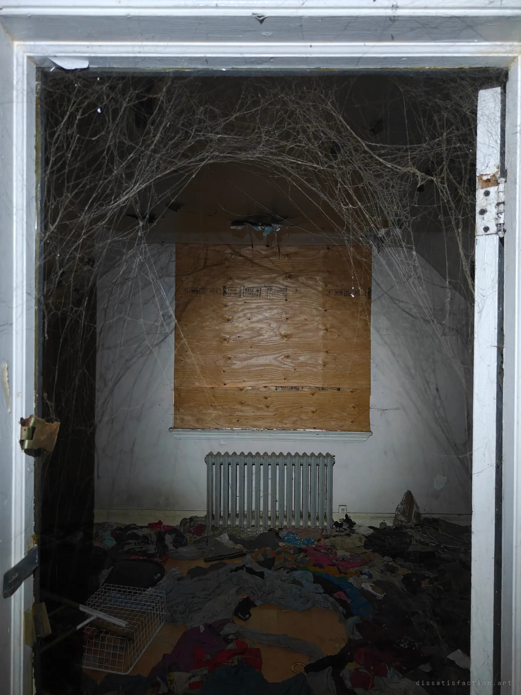 Looking through a doorway veiled in thick cobwebs into a bedroom with a boarded window above a radiator. Clothes are strewn ankle-deep across the floor.