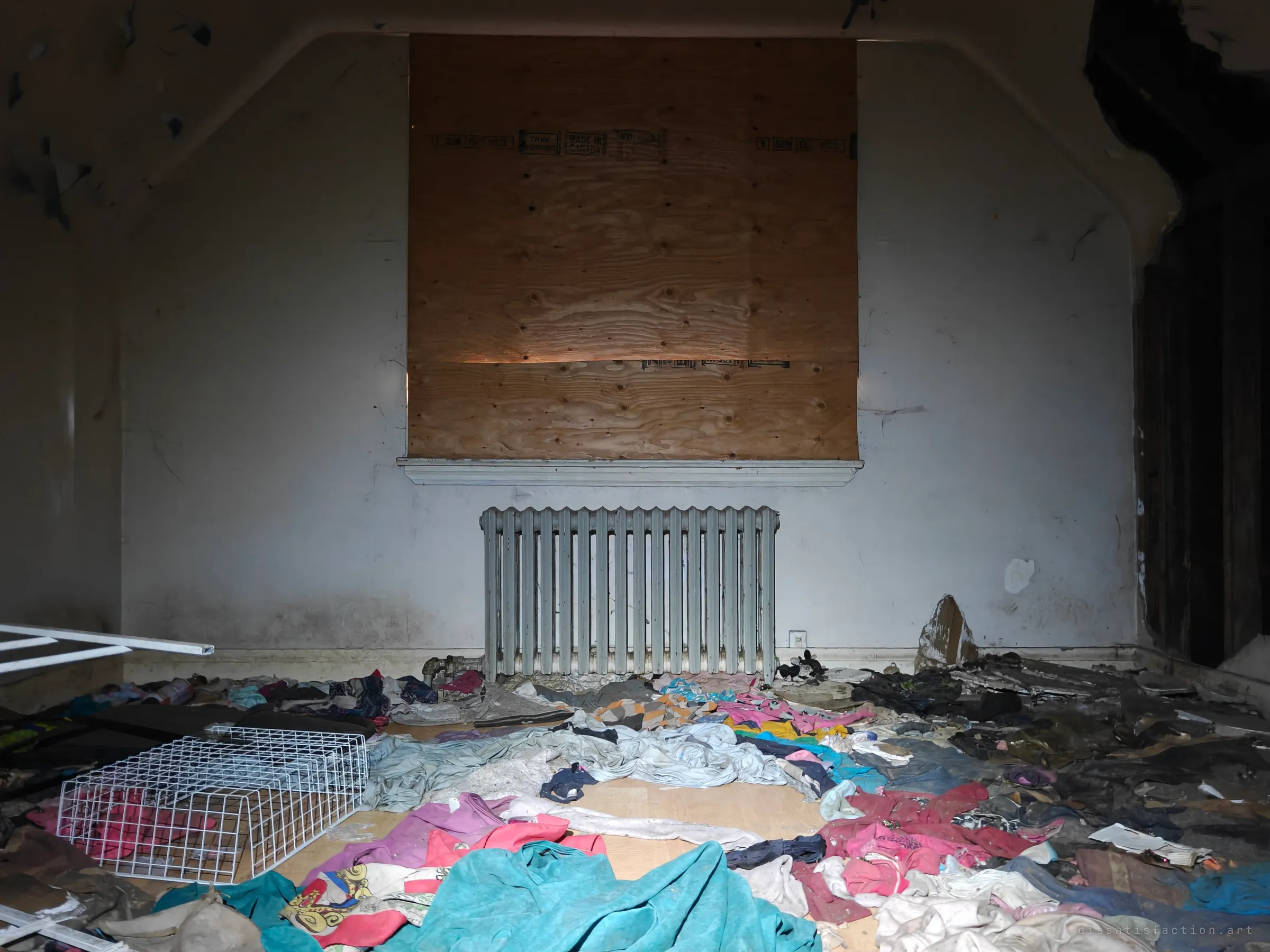 A boarded-up window above an old cast-iron radiator, with a floor carpeted in heaps of abandoned clothing. The plywood seal and scattered belongings show a home left behind in a hurry.