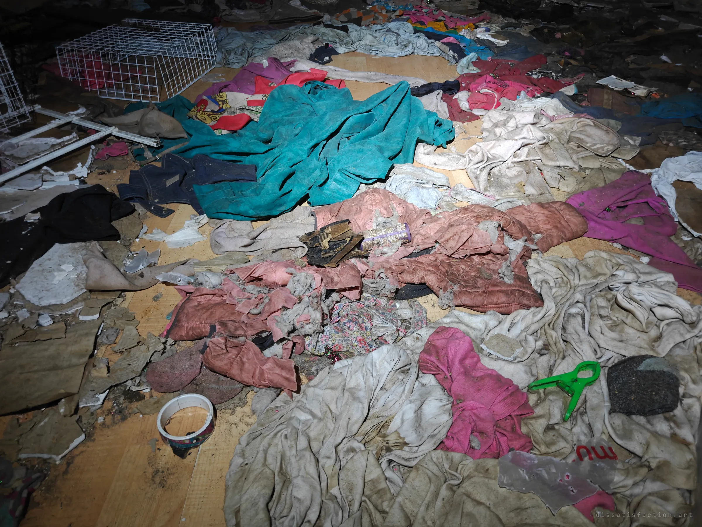 A close look at the floor of the same room: dusty jackets, shirts and a torn quilted coat piled among plaster fragments. The layers of everyday clothing make the abandonment feel personal.