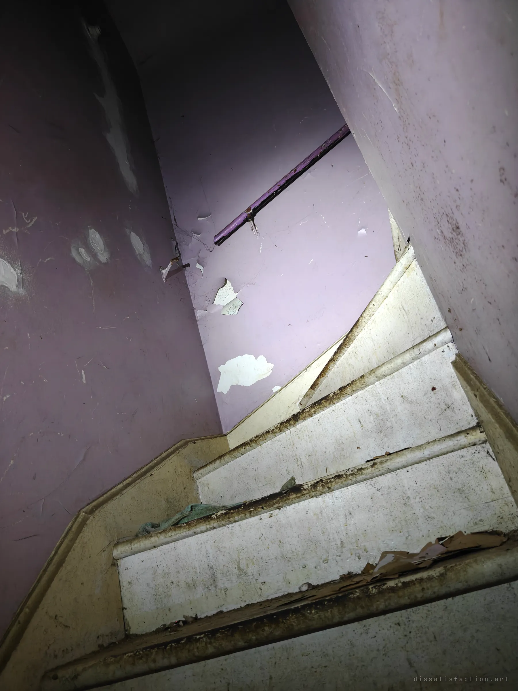 Looking up a narrow, steep staircase with lilac walls, peeling paint and a worn handrail. The tight turn and chipped white steps give the house a cramped, uneasy feel.