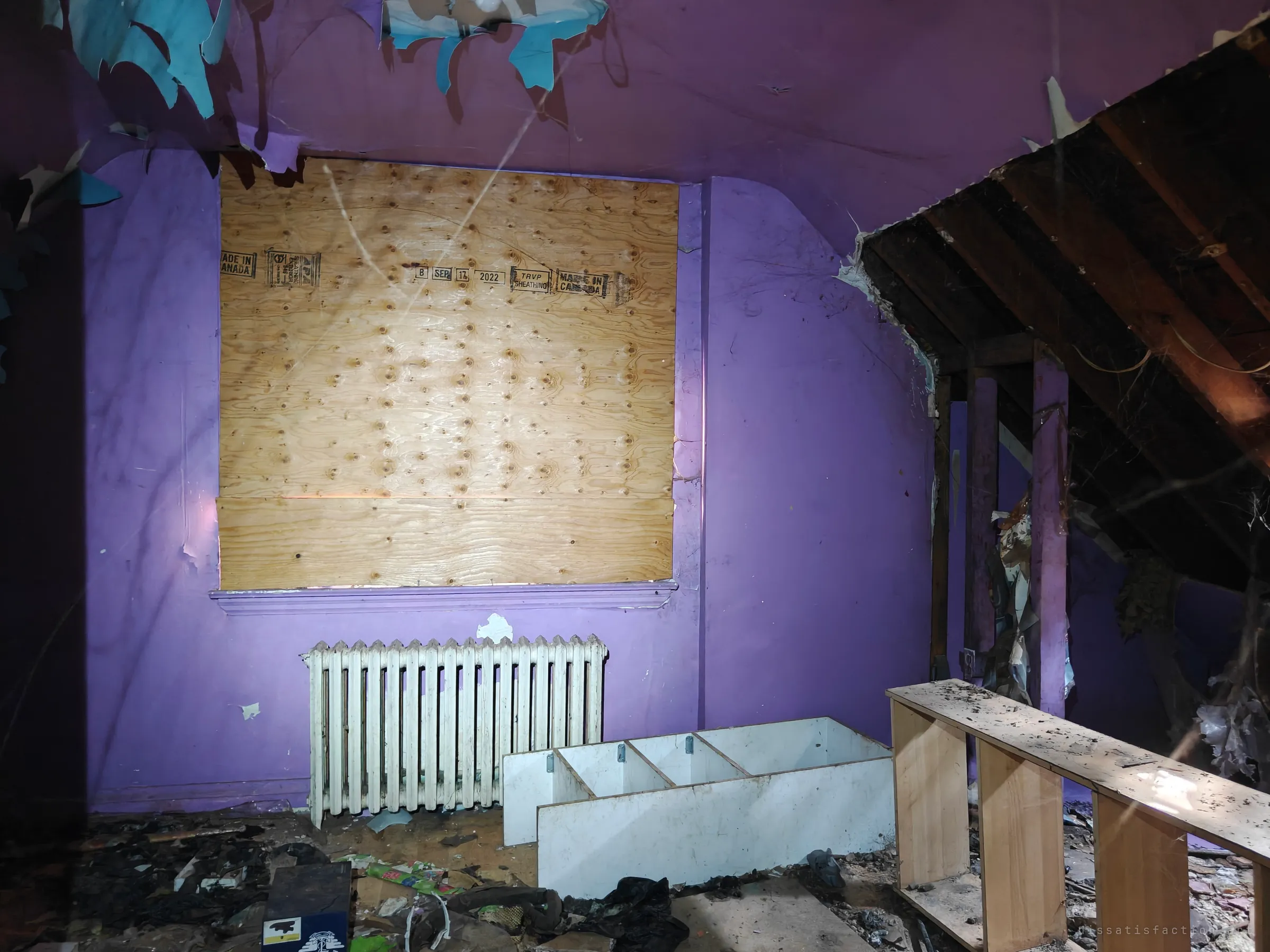 An attic bedroom painted violet, its window sealed with plywood above a white radiator, with the roof opened up to bare rafters on the right. Paper hangs from the ceiling like torn skin.