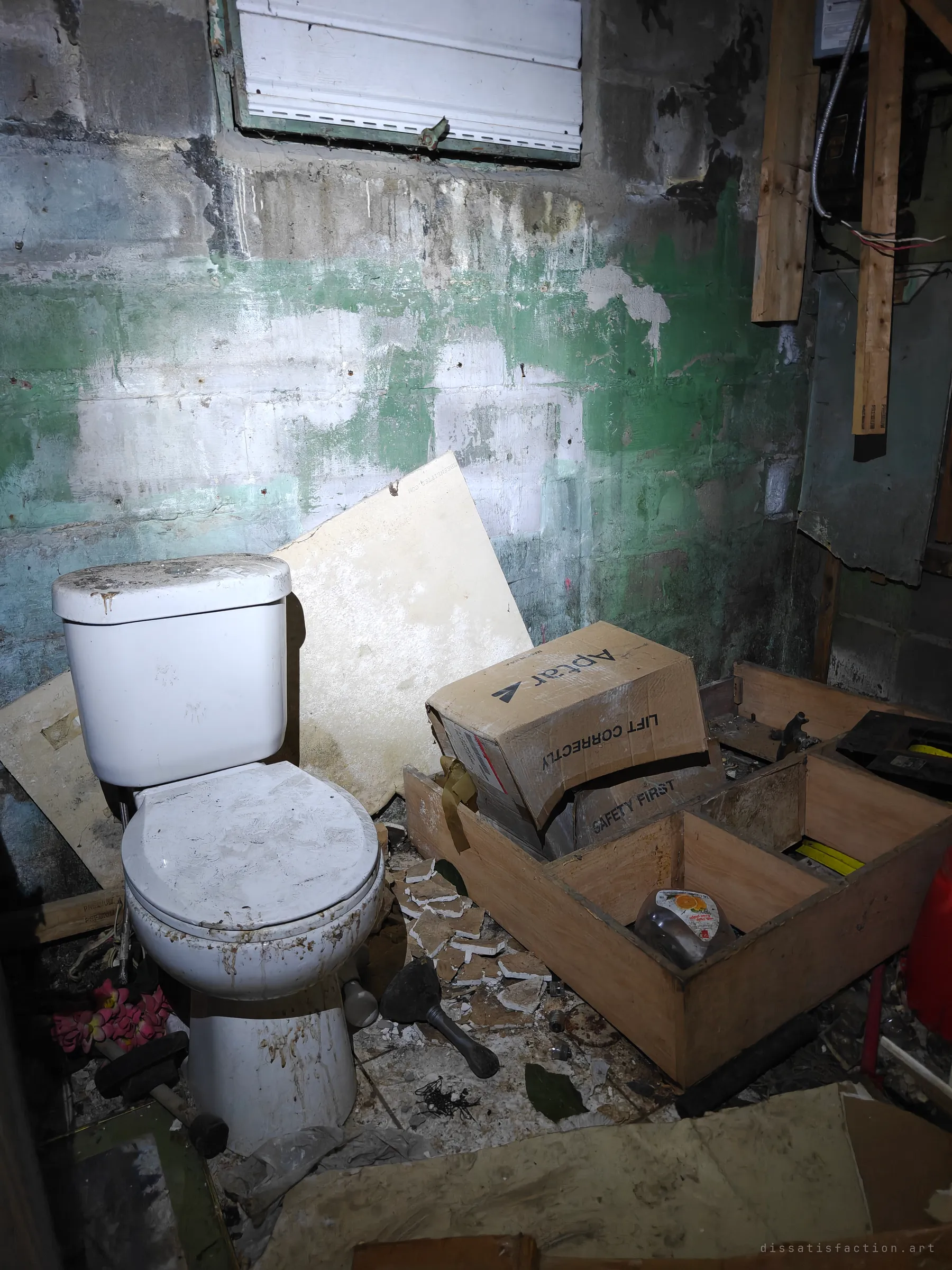 A basement toilet against a cinderblock wall streaked with green paint, surrounded by broken tiles, boxes and scrap wood. Exposed wiring and a covered window add to the decay.