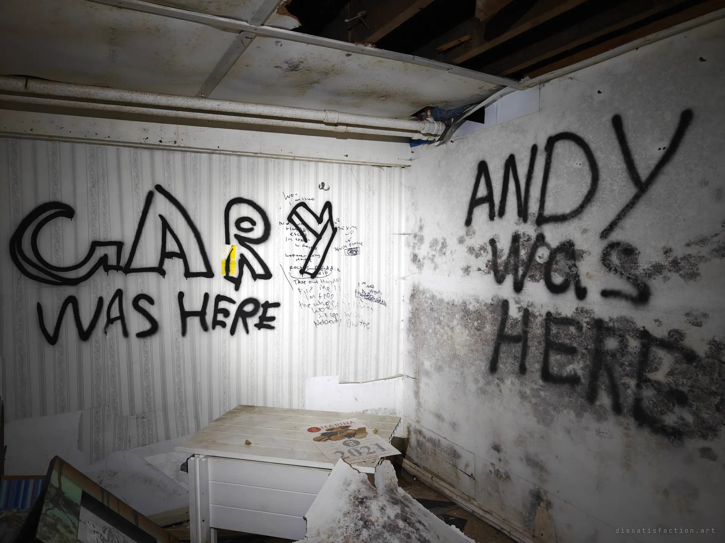 A basement room covered in large spray-painted "was here" tags and scrawled handwriting, with black mould spreading across the right wall. An old wall calendar lies on a small table.
