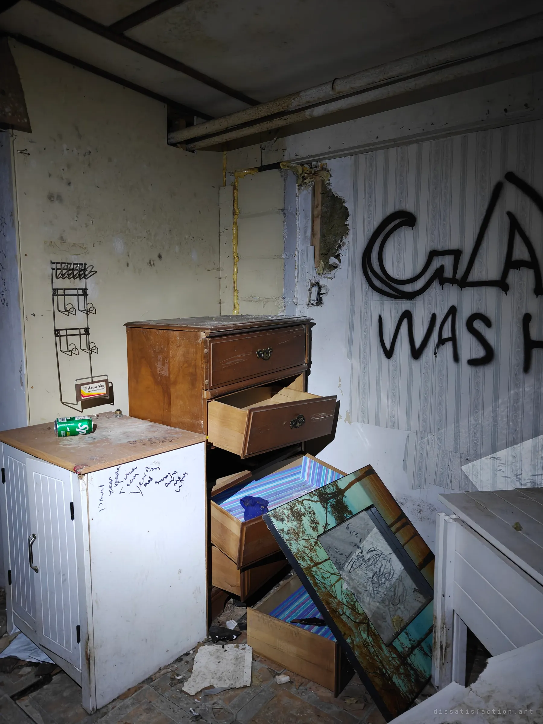 A corner of the same graffiti room: a dresser with drawers pulled out, a white cabinet scribbled with pen, and a leaning framed print. Pipes run along the low ceiling above.
