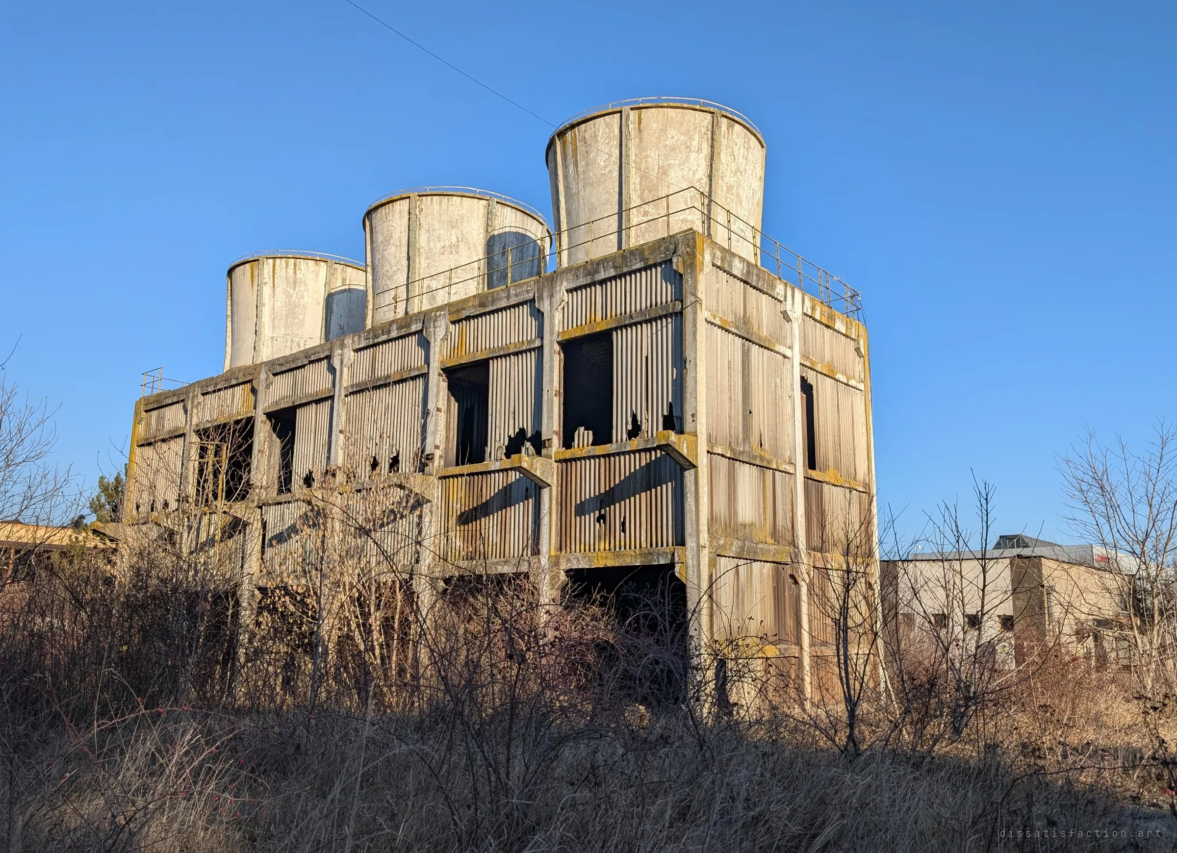A derelict concrete cooling block with three weathered round towers on its roof, sides clad in broken corrugated panels, rising above dry winter scrub under a clear blue sky. Sets the scale of the site.