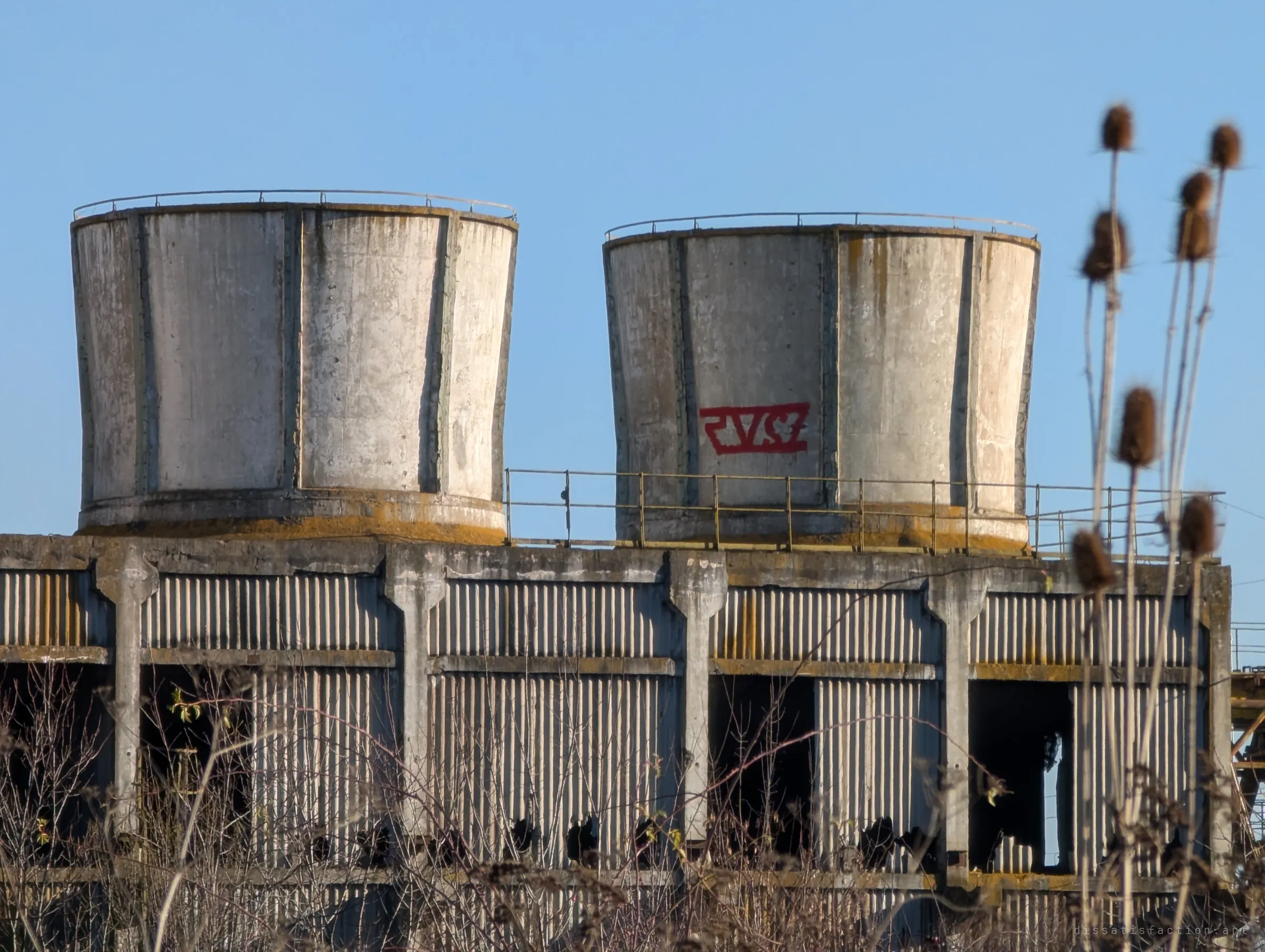 Telephoto view of two of the flared concrete cooling cells, one tagged with red graffiti, above rows of corrugated panels and dried teasel heads in the foreground.