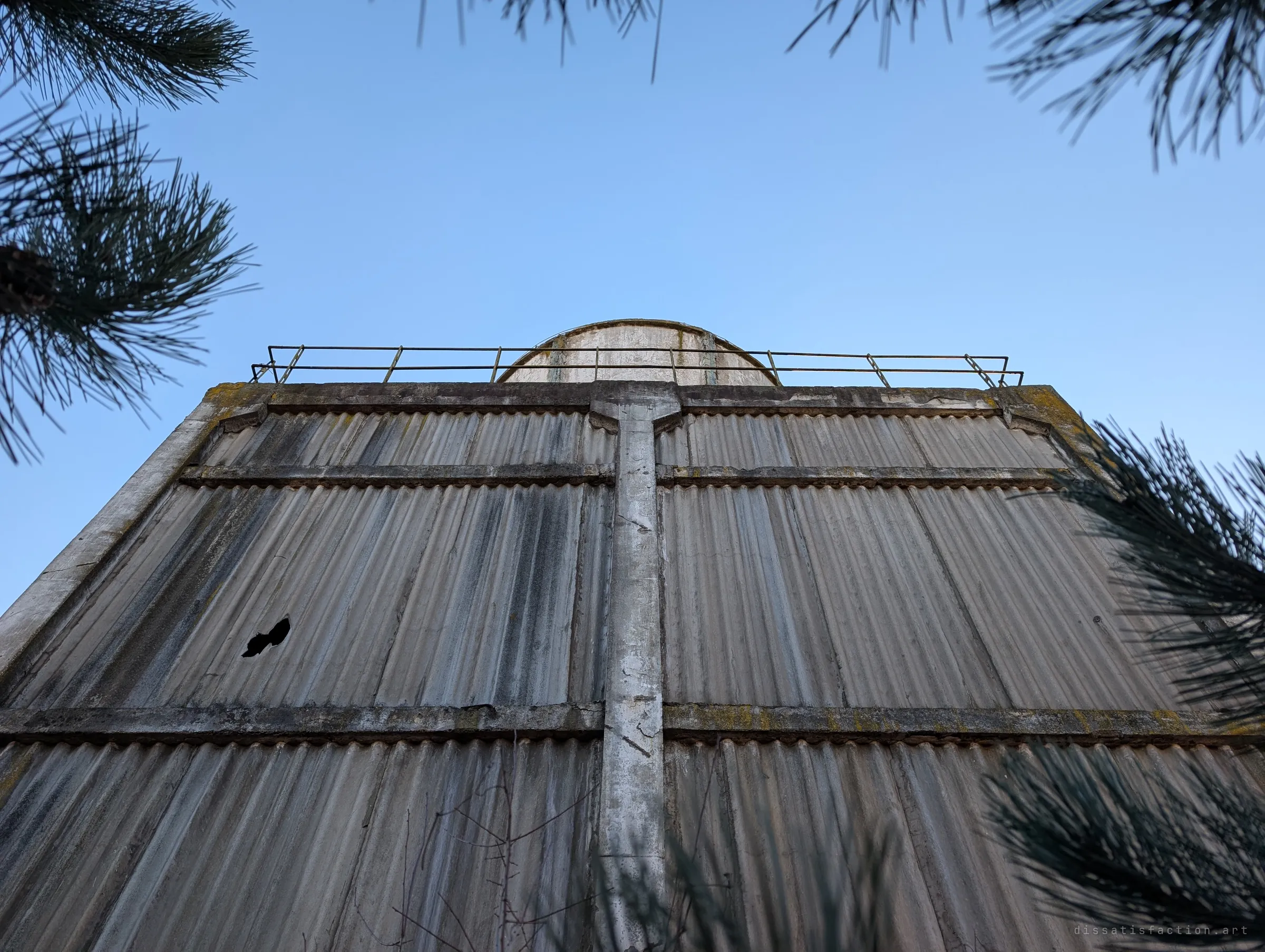 Looking straight up a corrugated wall of the cooling block toward the rim of one tower and its railing, framed by pine branches.