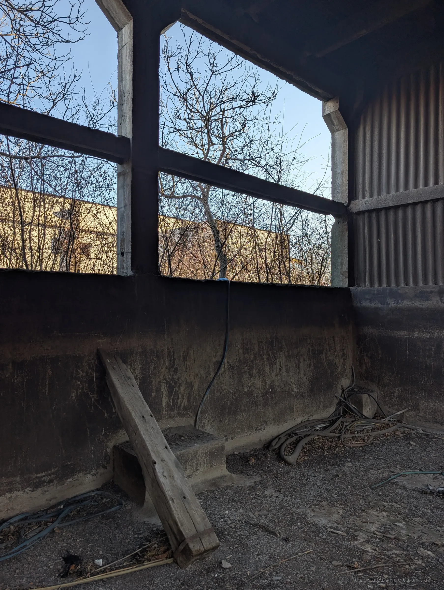 Inside the open base of the block: a soot-darkened basin wall, an old timber beam and tangled cables, with bare trees and a neighbouring building seen through the concrete frame.