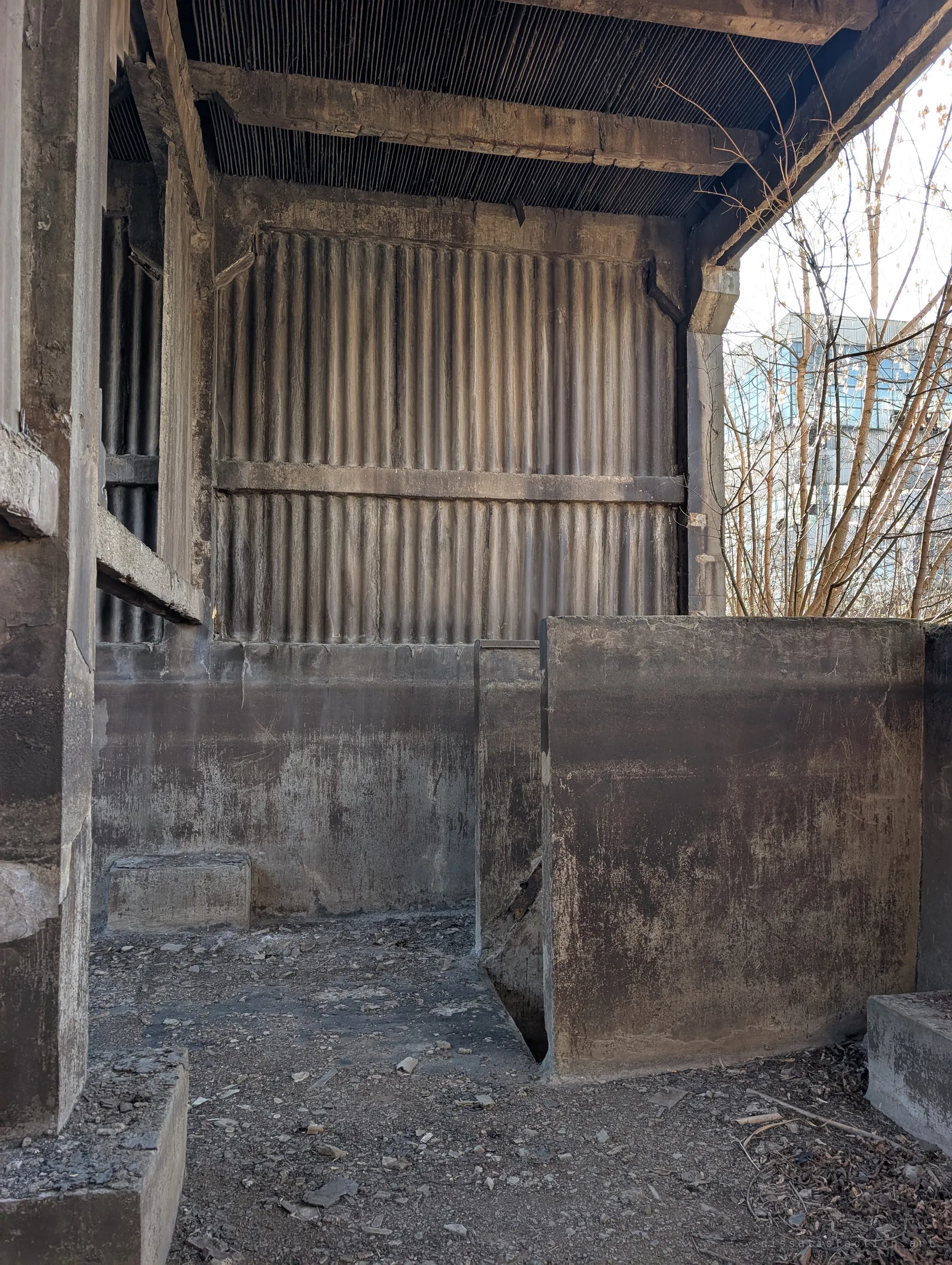 Blackened concrete basin walls and a narrow channel beneath the corrugated upper structure, the floor scattered with fallen debris.