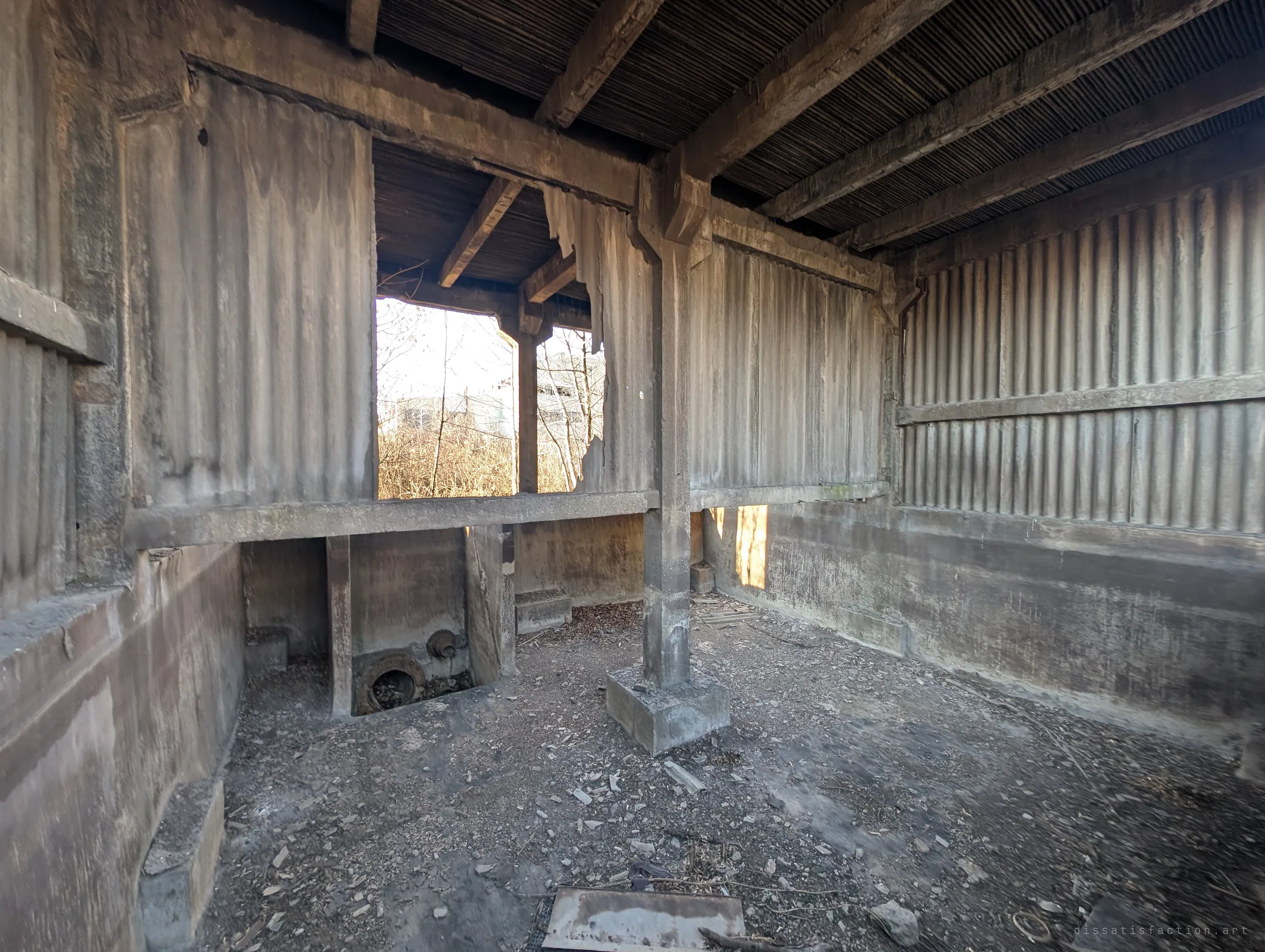 Wide view inside one empty cooling cell: corrugated walls, timber roof beams, a central pillar and a sunken pipe outlet, with light spilling through a torn opening.
