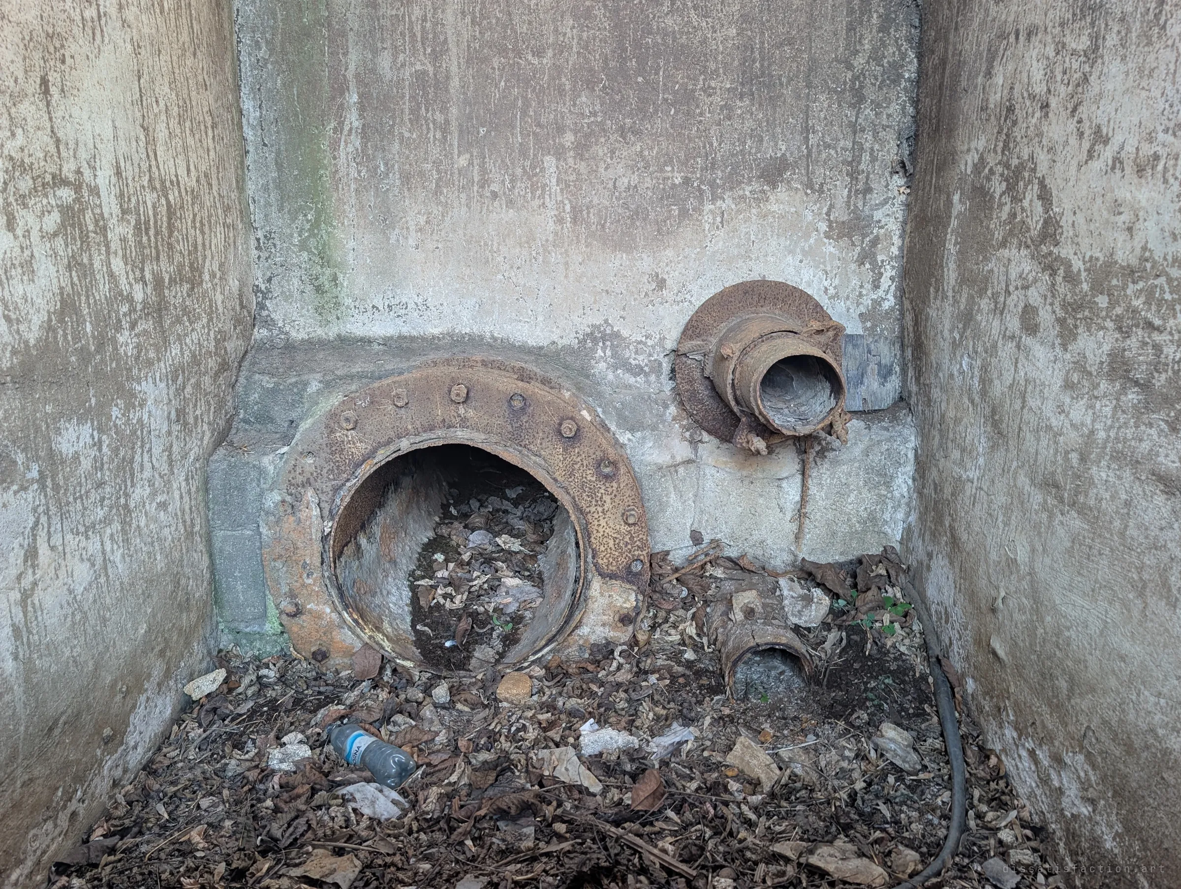 A corroded flanged outlet and a smaller broken pipe at the foot of a concrete sump, half buried in dry leaves and a discarded bottle.