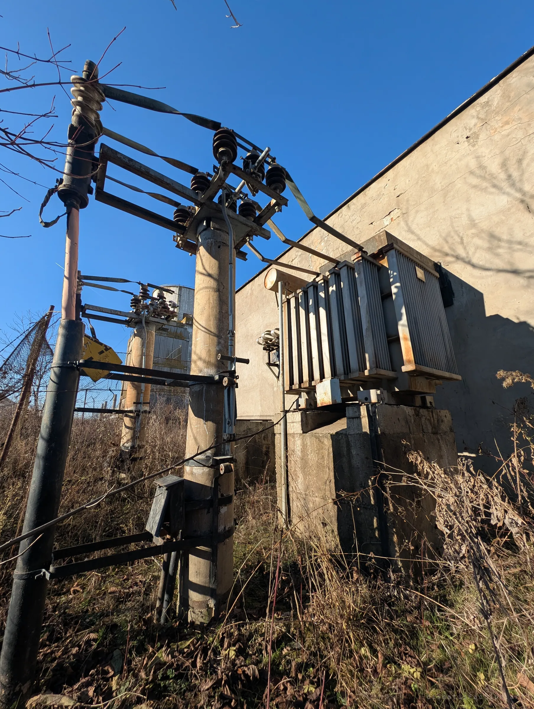 An old pole-mounted transformer and insulators beside a plain wall, a yellow danger sign hanging askew and a cooling cell behind the fence. Some of it may still be live.