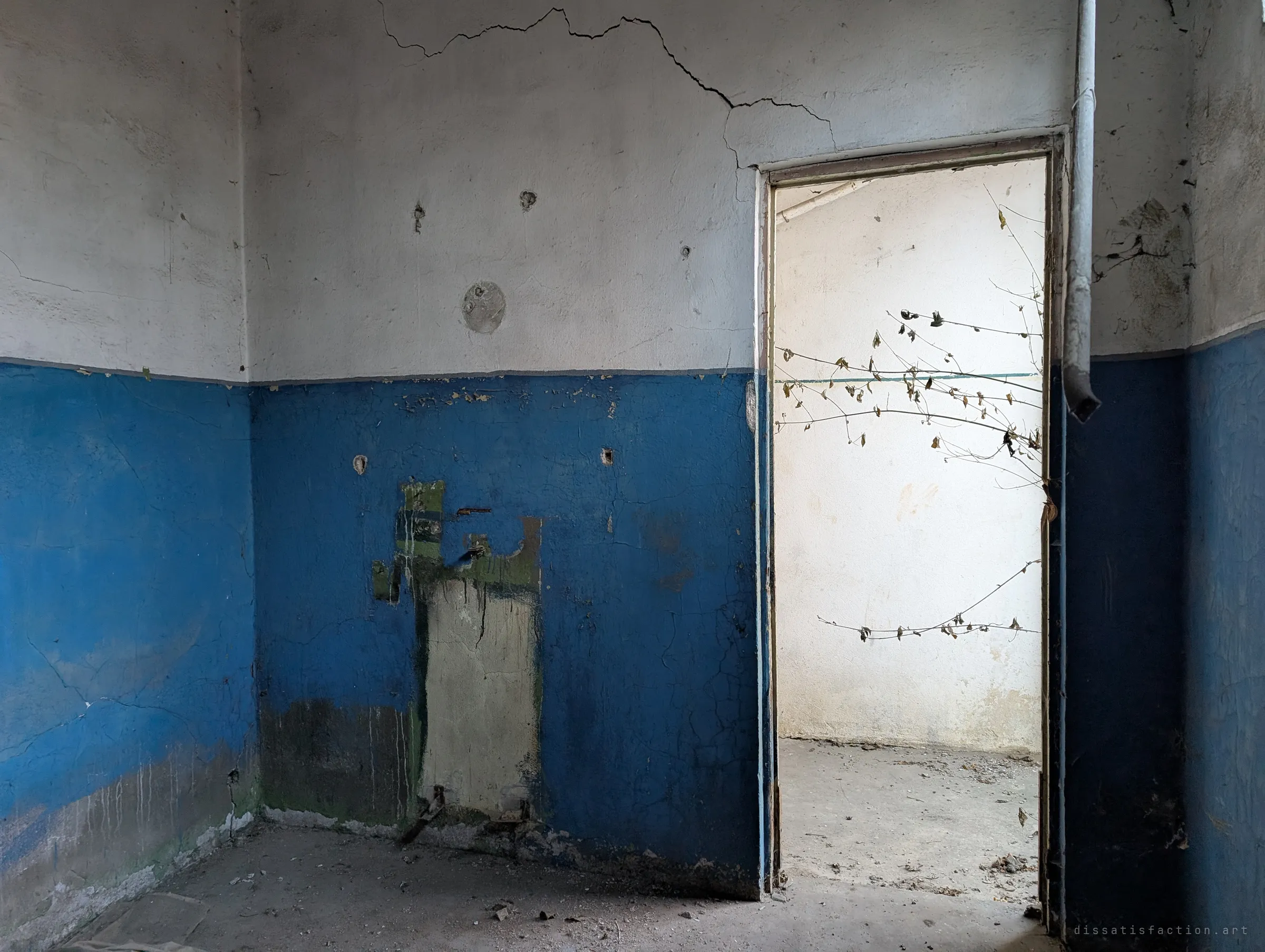 A cracked room painted half blue, half white, with a scar where a fixture was torn off and a doorway opening onto a bright empty room with a dead plant hanging in it.