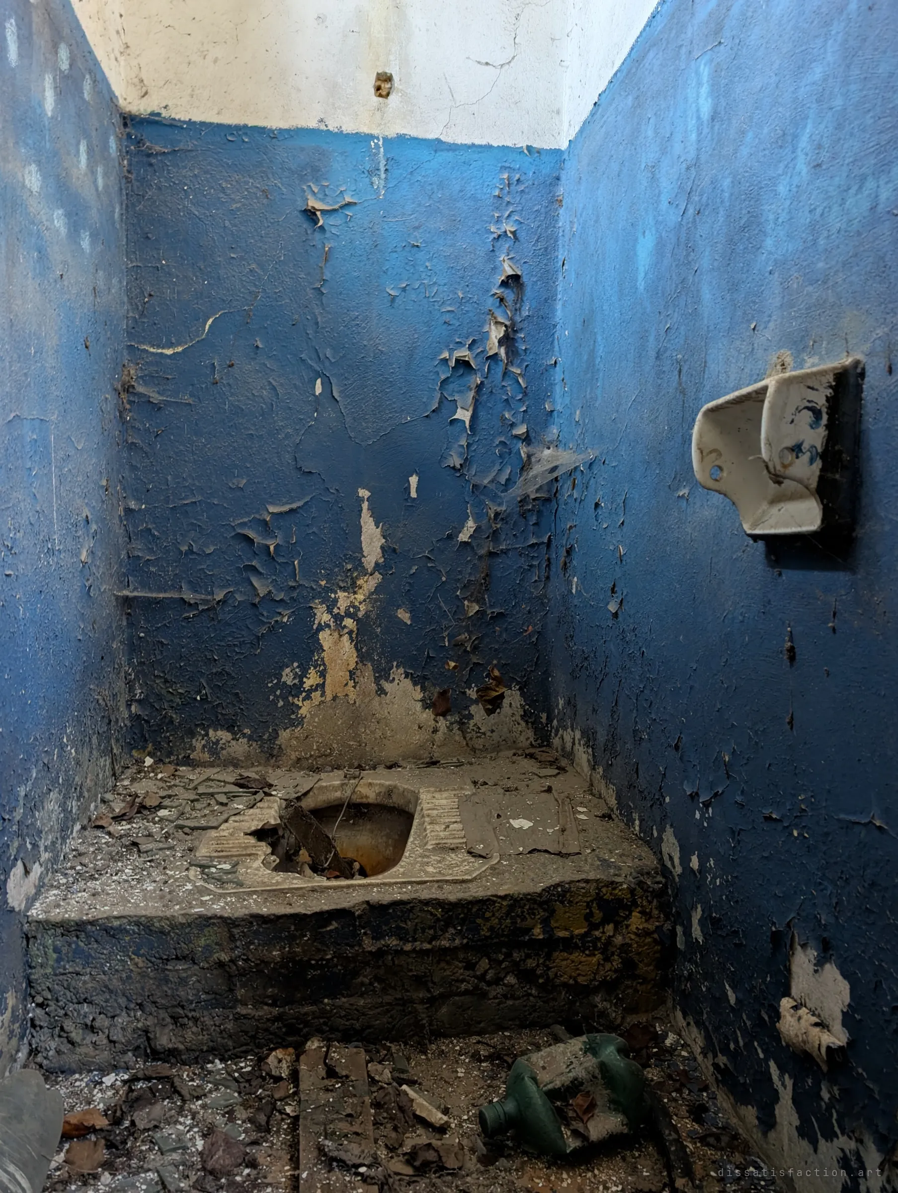 A cramped blue washroom stall with peeling paint, a broken squat toilet and a stained ceramic soap dish, glass and a green bottle on the floor.