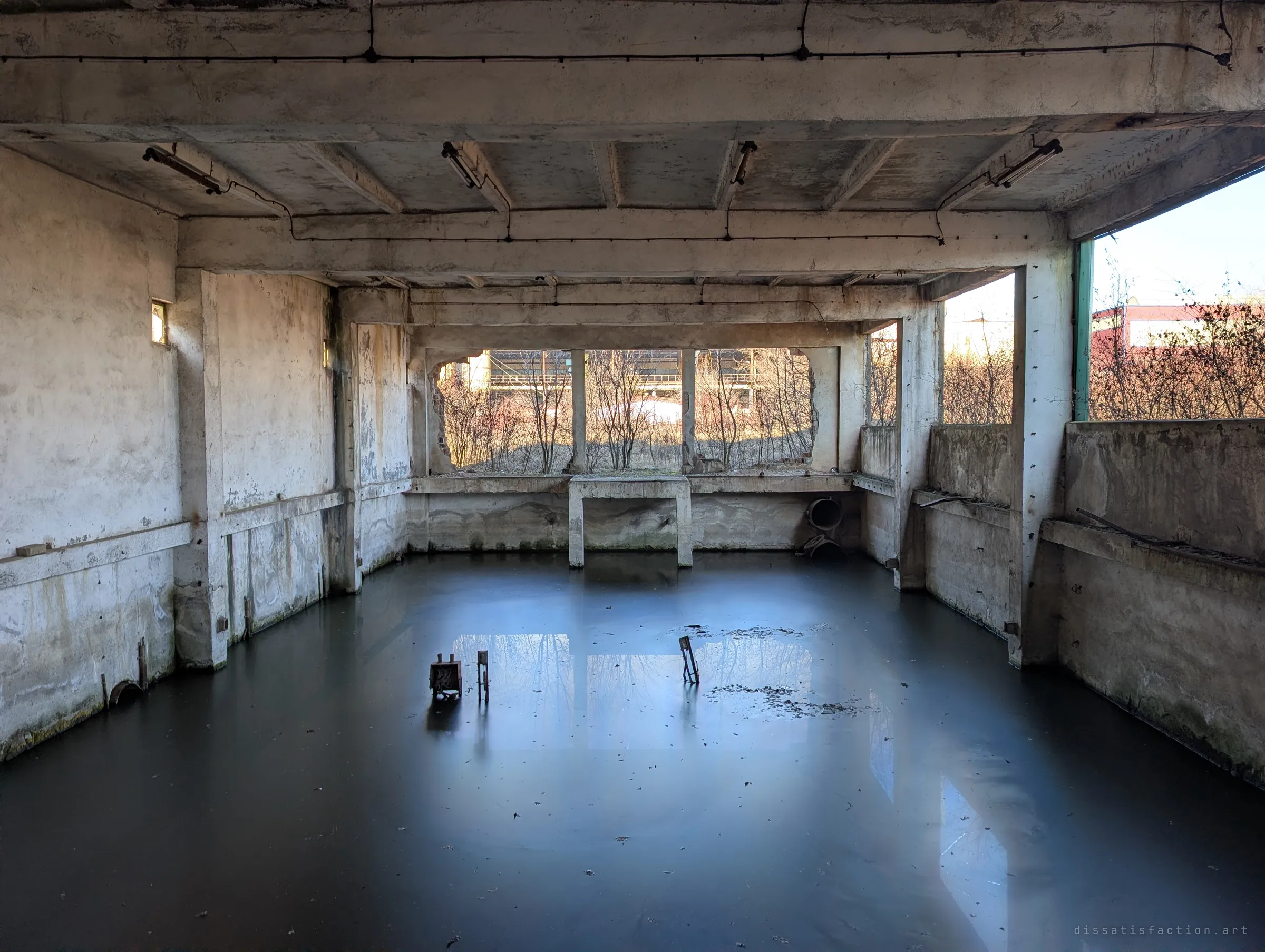 A flooded ground-floor hall under a ribbed concrete ceiling, the black water perfectly still and mirroring the sunlit openings. A rusted electrical box stands alone in the middle, the only thing left above the surface.