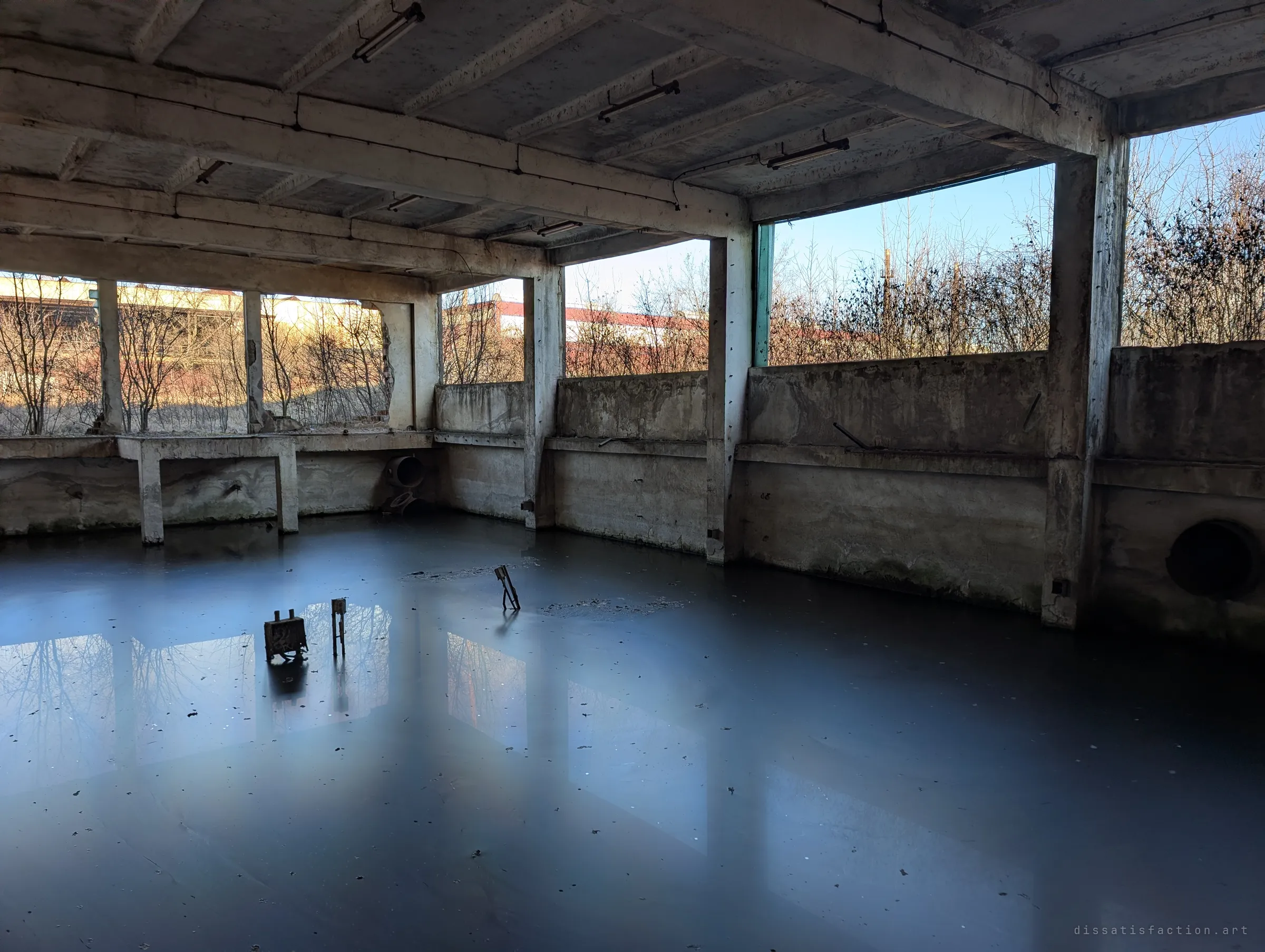 The same flooded hall seen from a corner, low winter sun pouring through the open wall and laying pale reflections across the dark water. Rusted fittings poke up from the surface near a round drain opening.