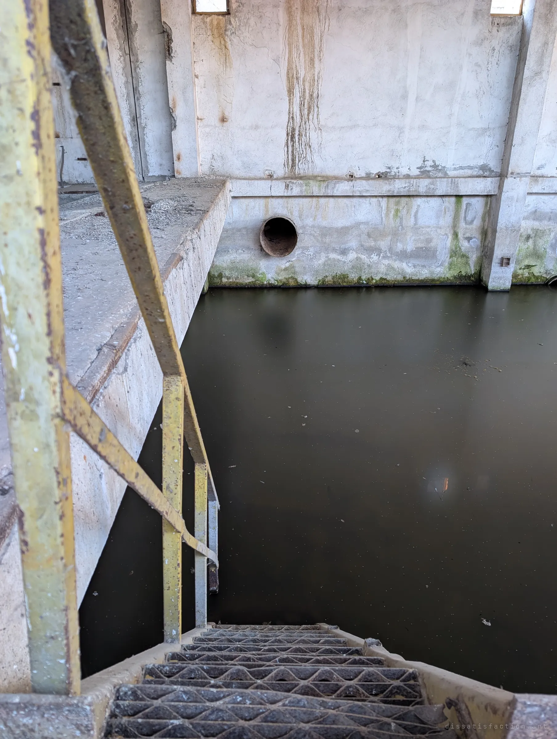 Looking down a rusty yellow stair whose treads disappear into murky standing water, with a stained concrete wall and outlet pipe beyond. The steps simply end in the flood, with no way of telling the depth.