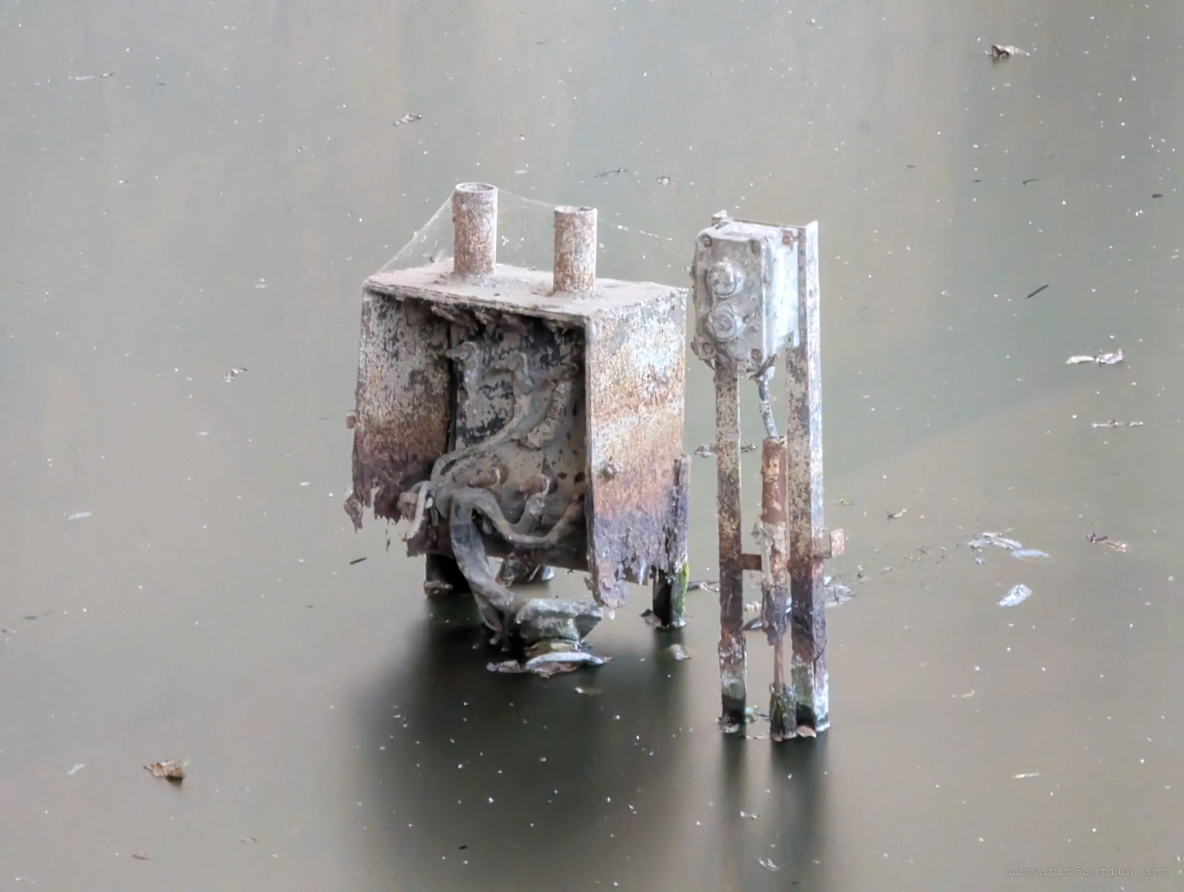 Close-up of a corroded electrical box and a switch post standing in the flat, still water, cobwebs strung between its pipes. Its innards spill out in rusted tangles just above the surface.