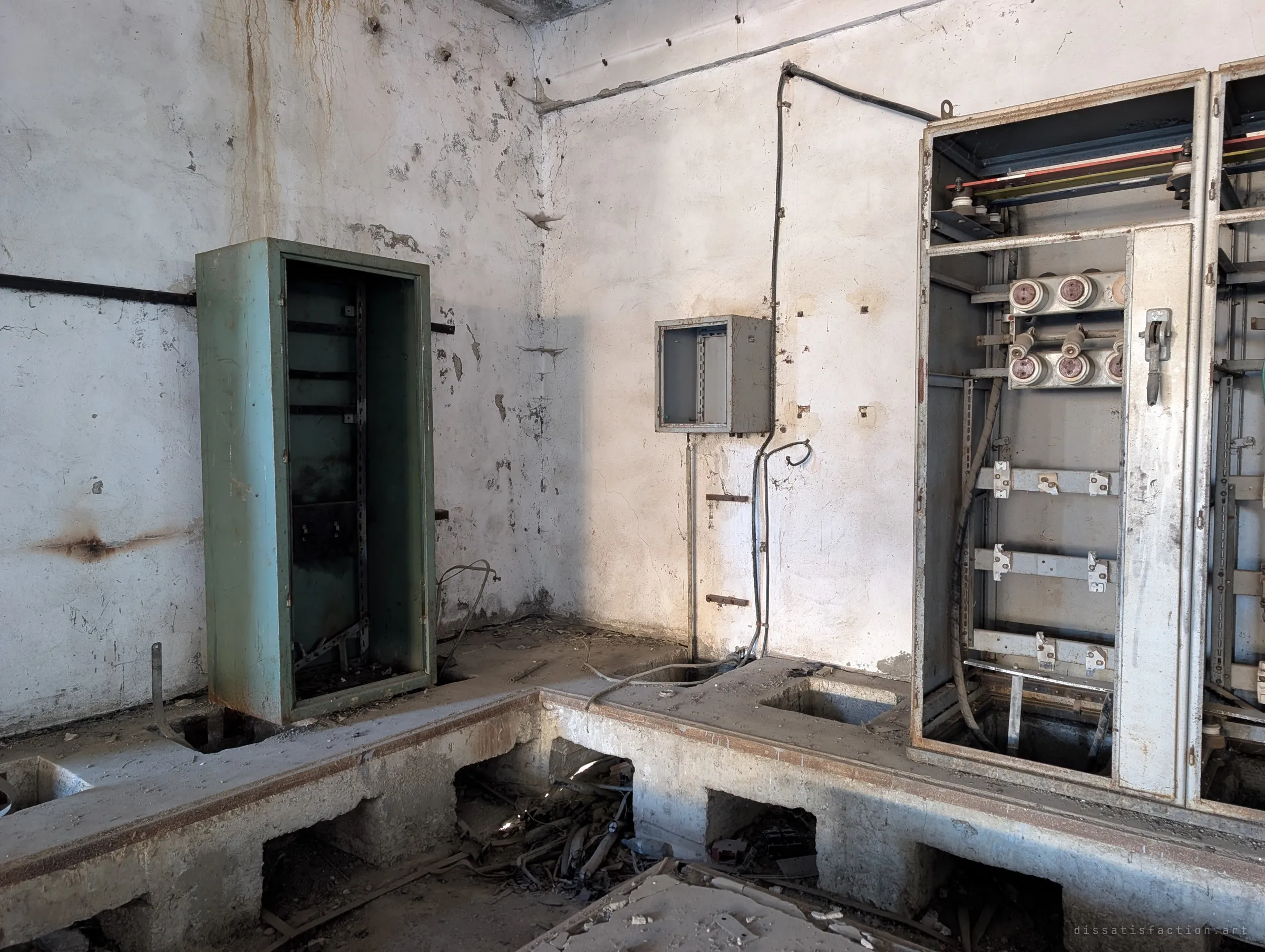 A stripped switch room with an open green cabinet, a small empty wall box and a gutted grey switchboard on a raised concrete plinth. Cable trenches below the plinth are choked with torn wiring.