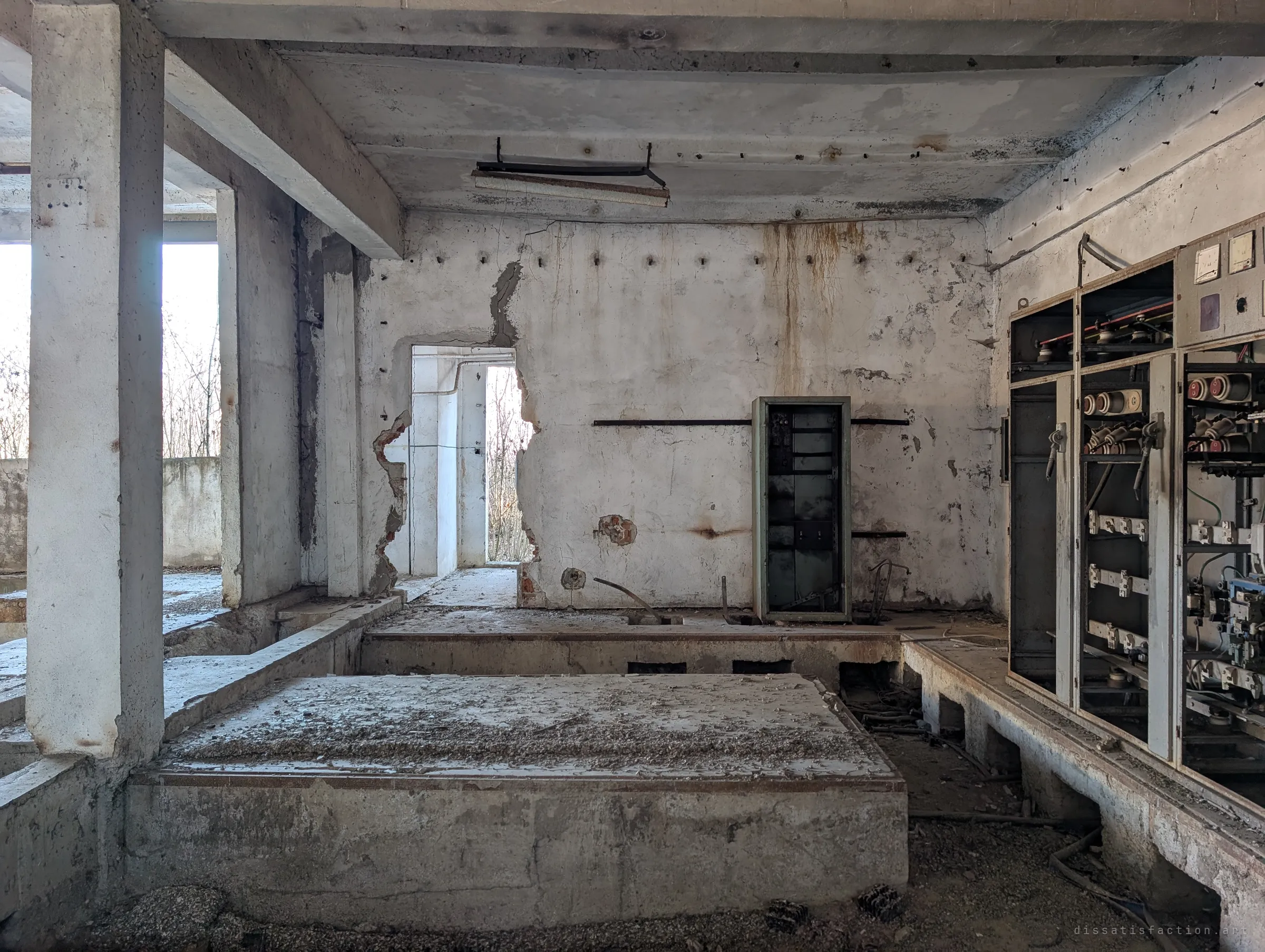The dry switch room, with a row of gutted switchboards on the right and a broken doorway leading through to daylight at the back. Dust and rubble cover the raised concrete bases where equipment once stood.