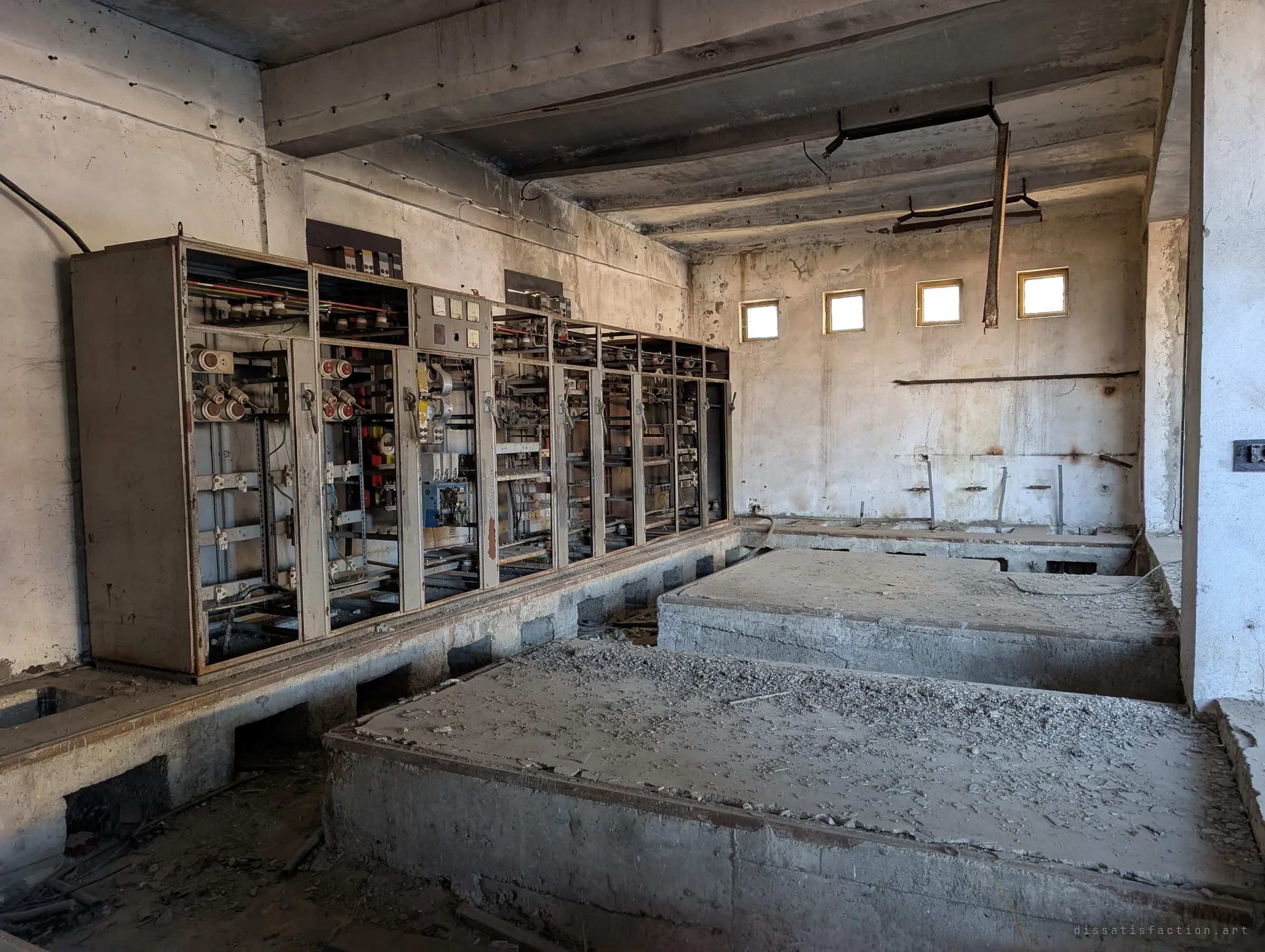 A long bank of open switchgear cabinets along the wall, dials and ceramic fuses still in place, facing empty concrete plinths. Small square windows high on the back wall light the dusty room.