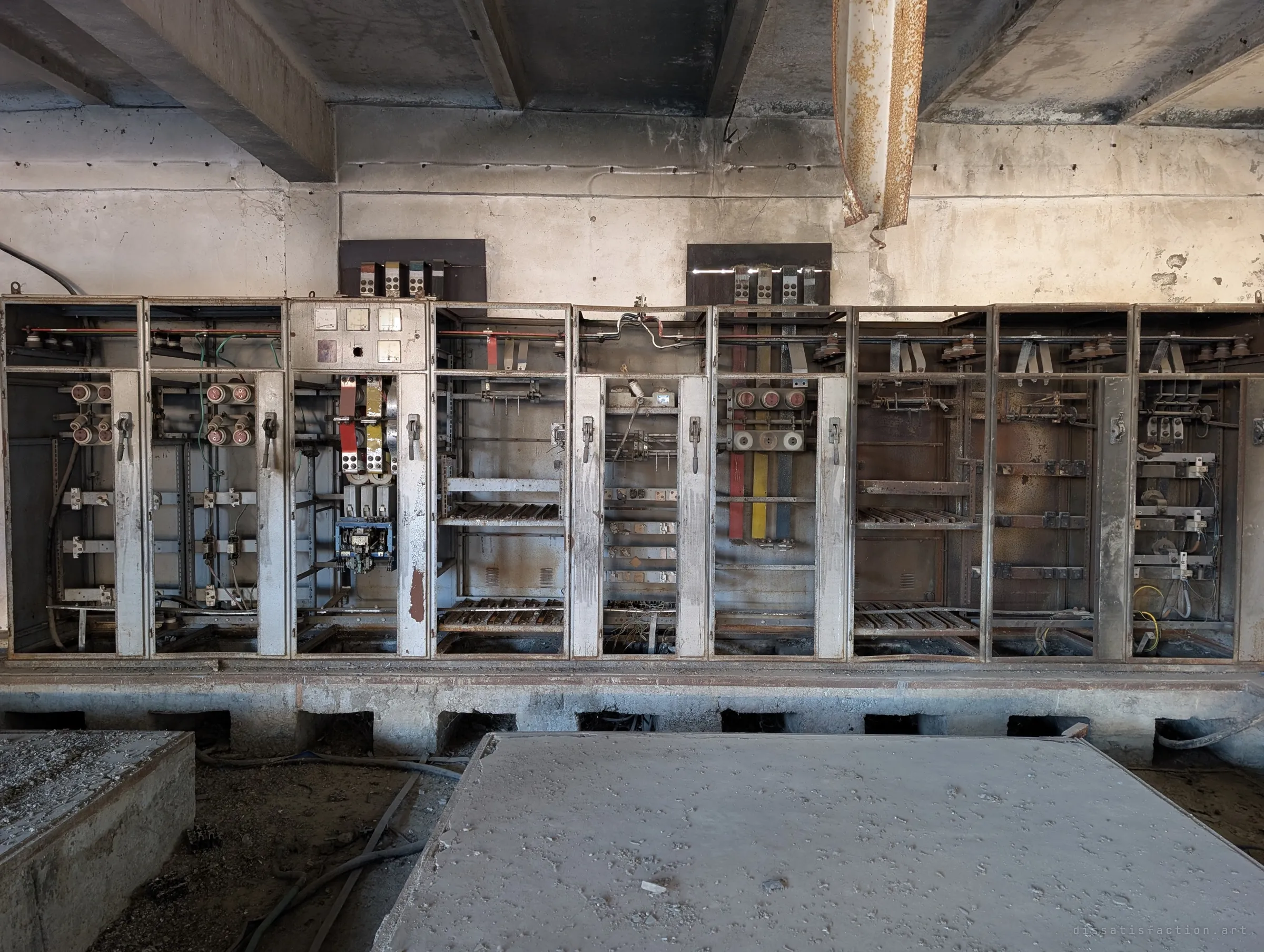 The full row of switchgear cabinets seen straight on, doors gone and busbars exposed in red, yellow and blue. A rusted pipe hangs loose from the ceiling above them.