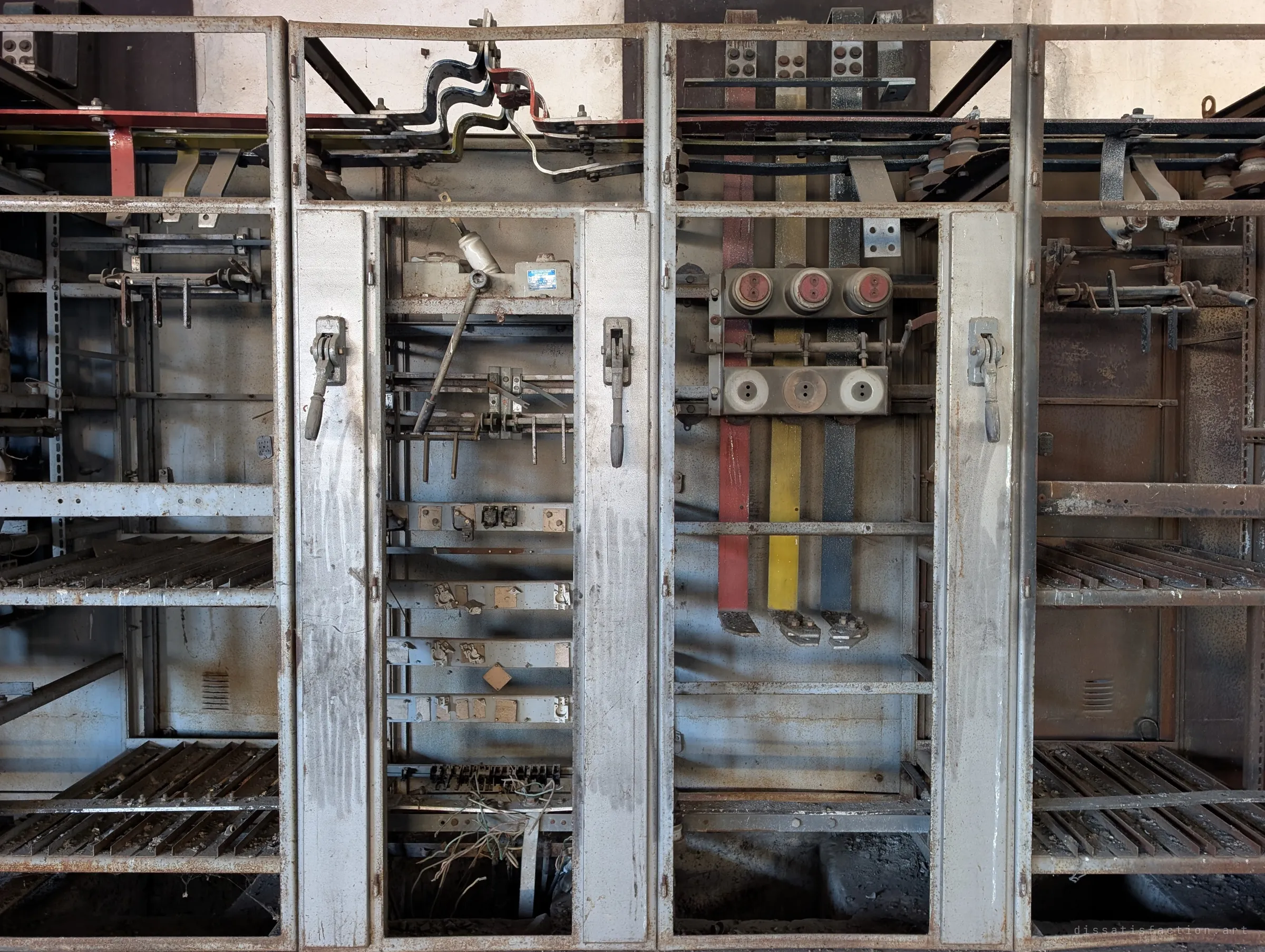 A tighter look into two switchgear bays: lever handles on the frames, empty rails, and three painted busbars in red, yellow and blue under a set of ceramic fuses. Decades of rust and dust coat every surface.