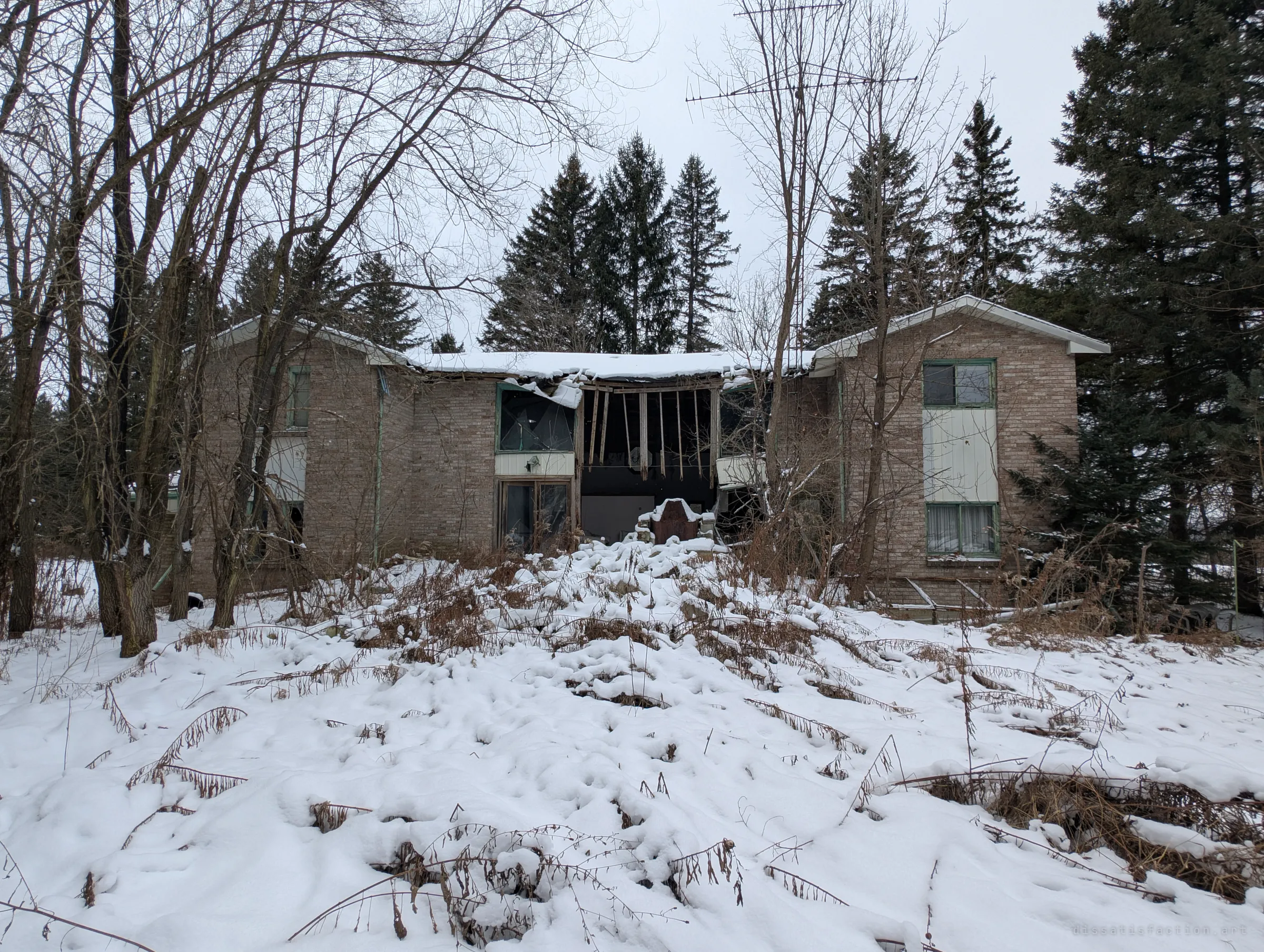 A two-storey brick house half buried in snow, its middle section caved in between two intact gables. Bare trees and spruces close in around it, and an old TV antenna rises above the roof.