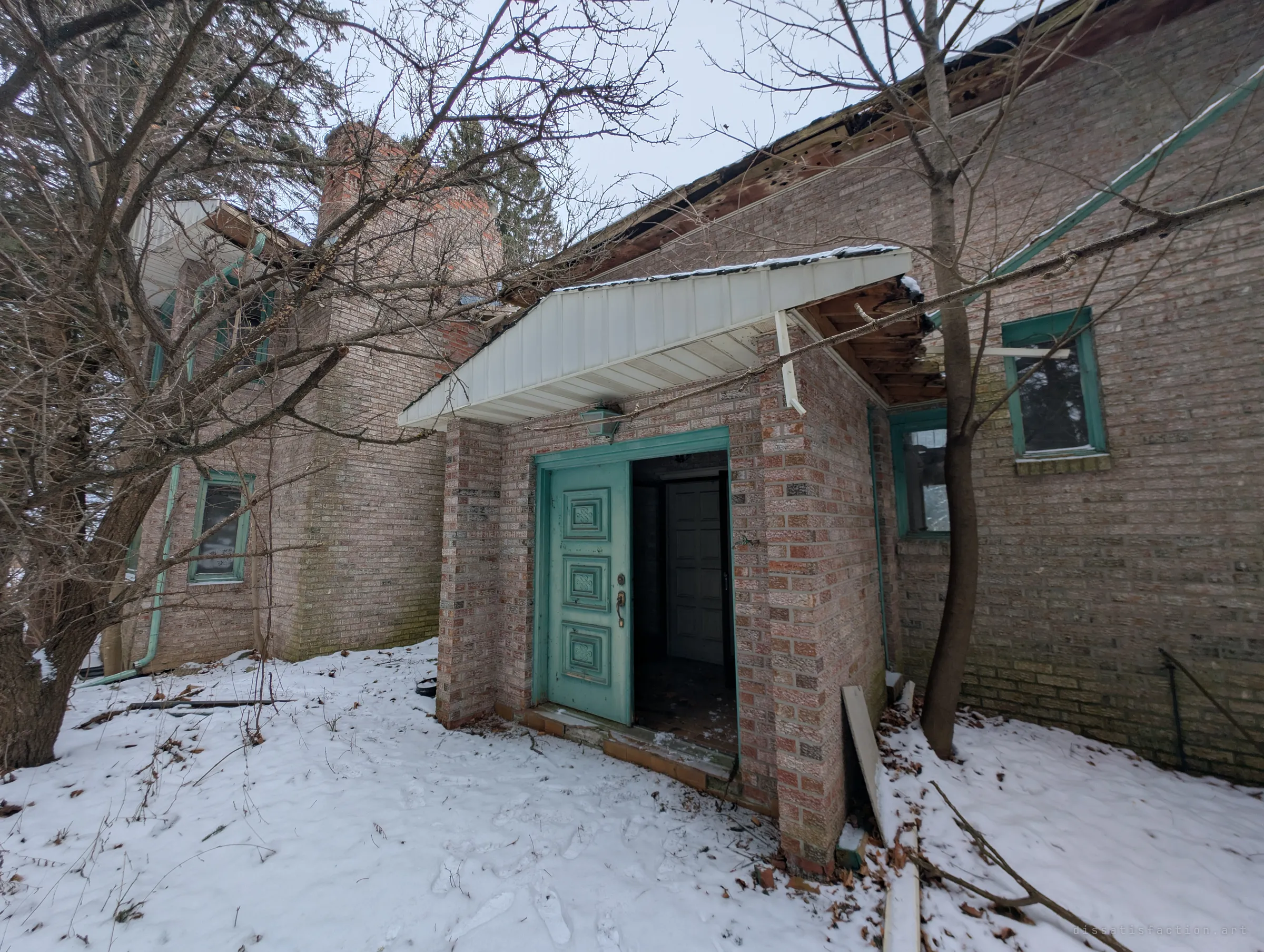 A small brick entry porch with a panelled teal front door standing open into darkness. The roof overhang above it is rotting away, and footprints lead through the snow.