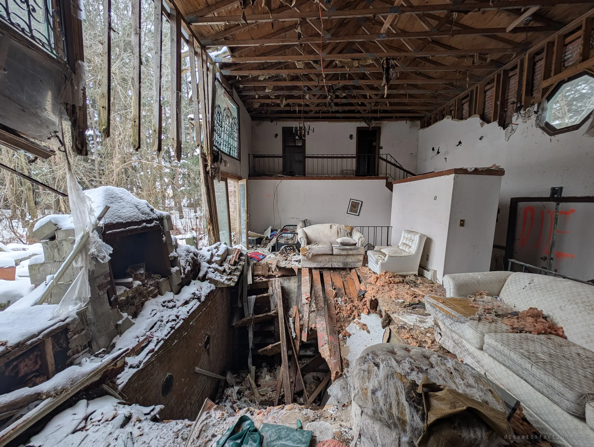 A double-height living room with the ceiling stripped to bare rafters. The floor has collapsed into the basement beside a snow-covered fireplace, while white sofas and a balcony landing are left behind. 'DIE' is spray-painted on a door.