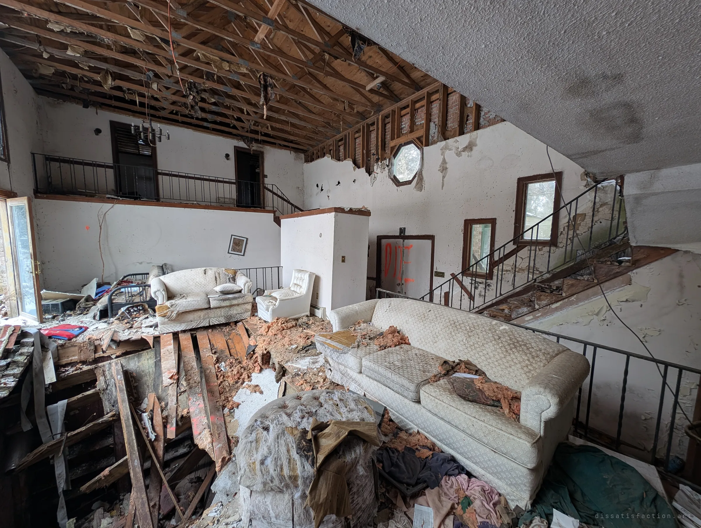 A wider view of the same living room: exposed roof trusses, an octagonal window, and a staircase whose treads are peeling away. Floorboards have given way next to a white sofa covered in insulation.