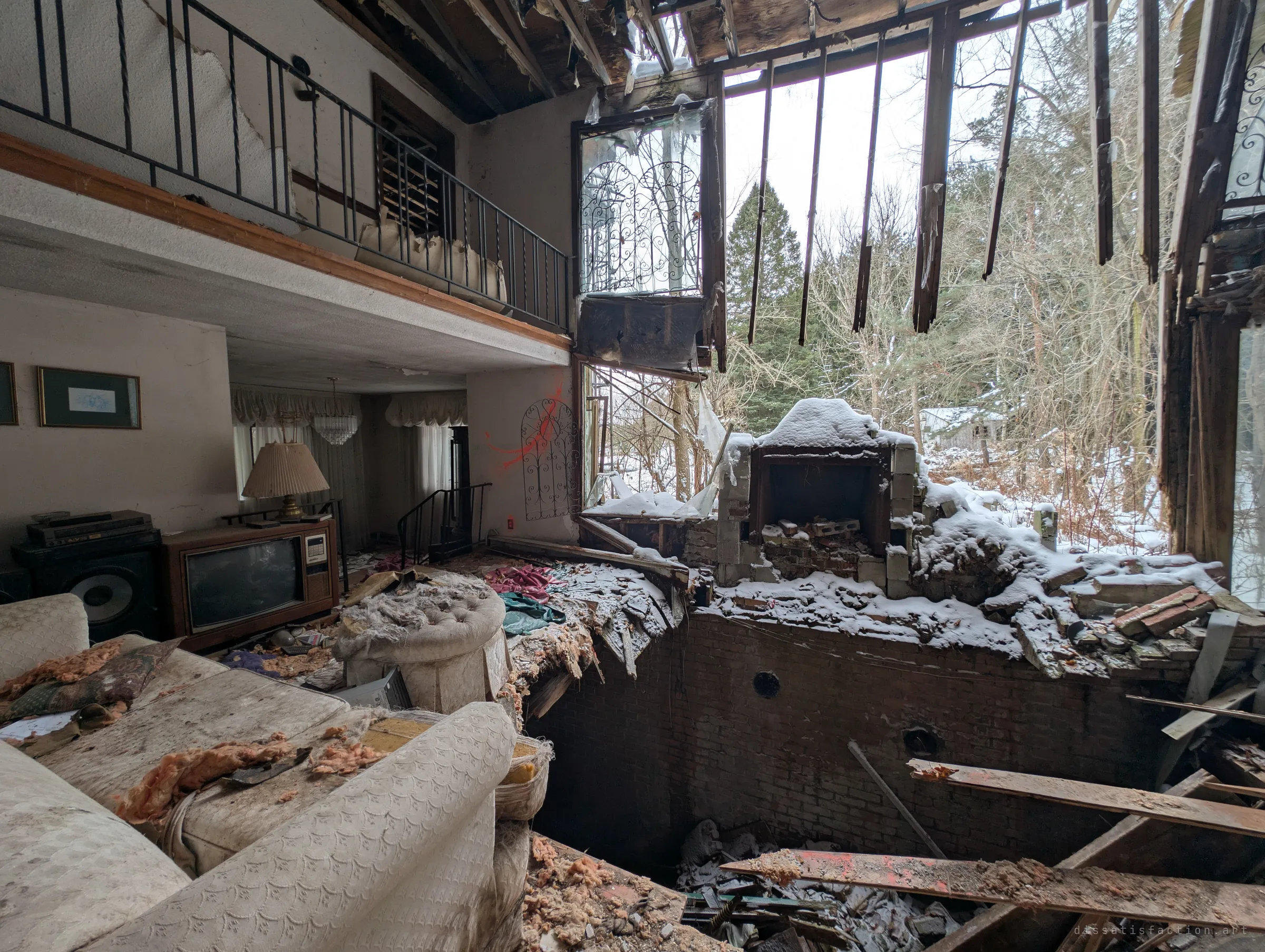 The glass wall has fallen away, so the living room opens straight onto the snowy woods. A brick fireplace stands on the edge of a pit where the floor dropped into the basement, and an old television and a table lamp still wait under the landing.