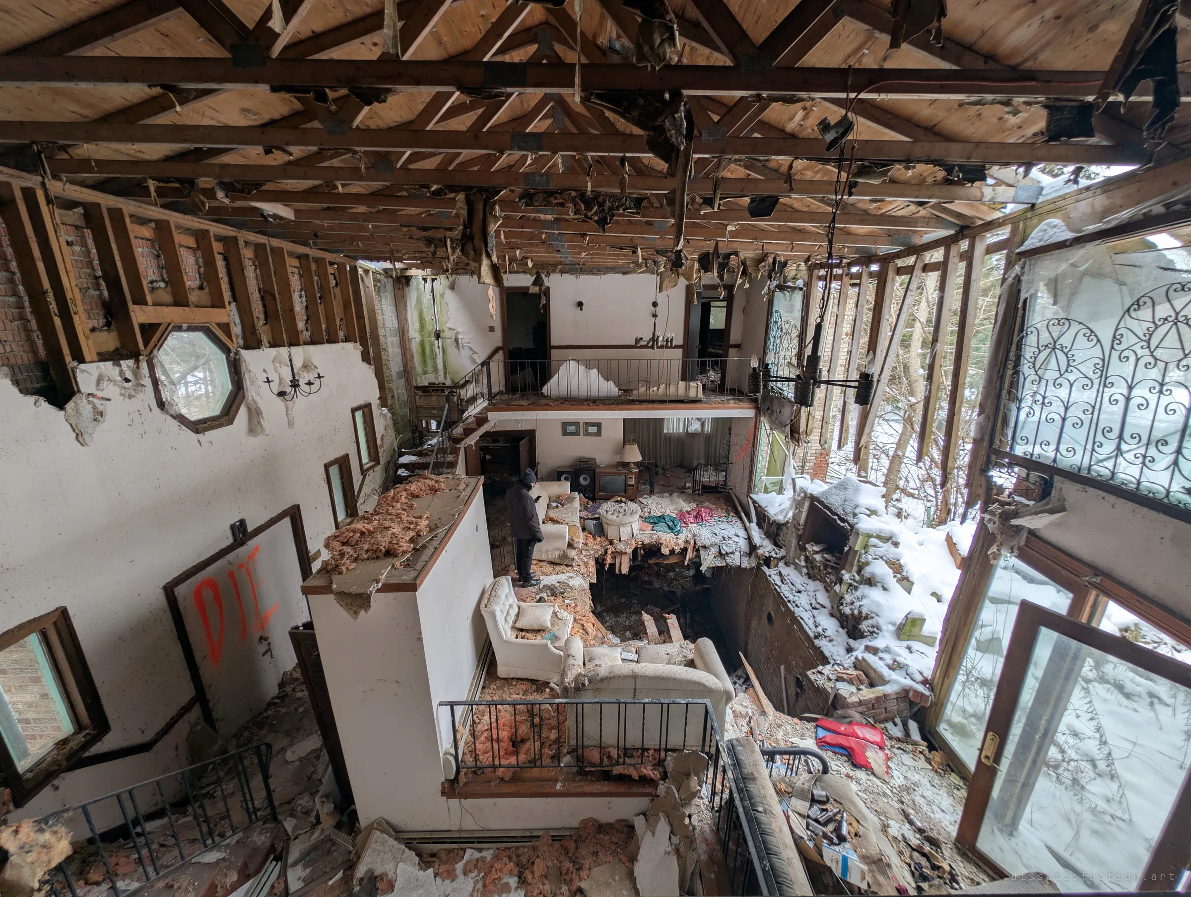 Looking down from the upper landing across the whole ruined great room. A lone explorer stands near the sofas, showing the scale of the gaping floor, the bare roof and the snow blowing in through the broken windows.