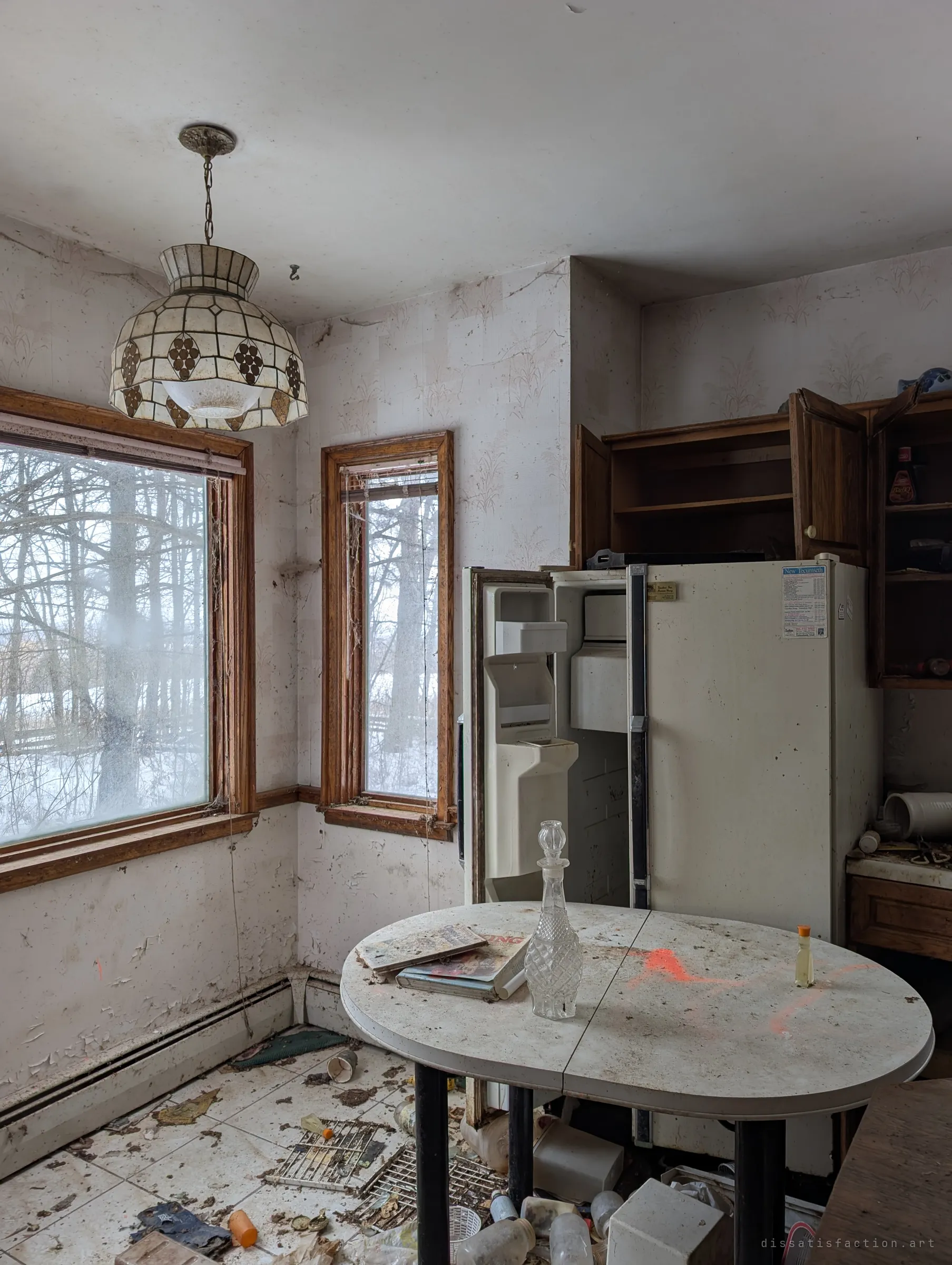 The breakfast nook beside the kitchen, with a leaded-glass pendant lamp hanging over an oval table. Winter light comes through tall windows onto the peeling walls and scattered bottles.